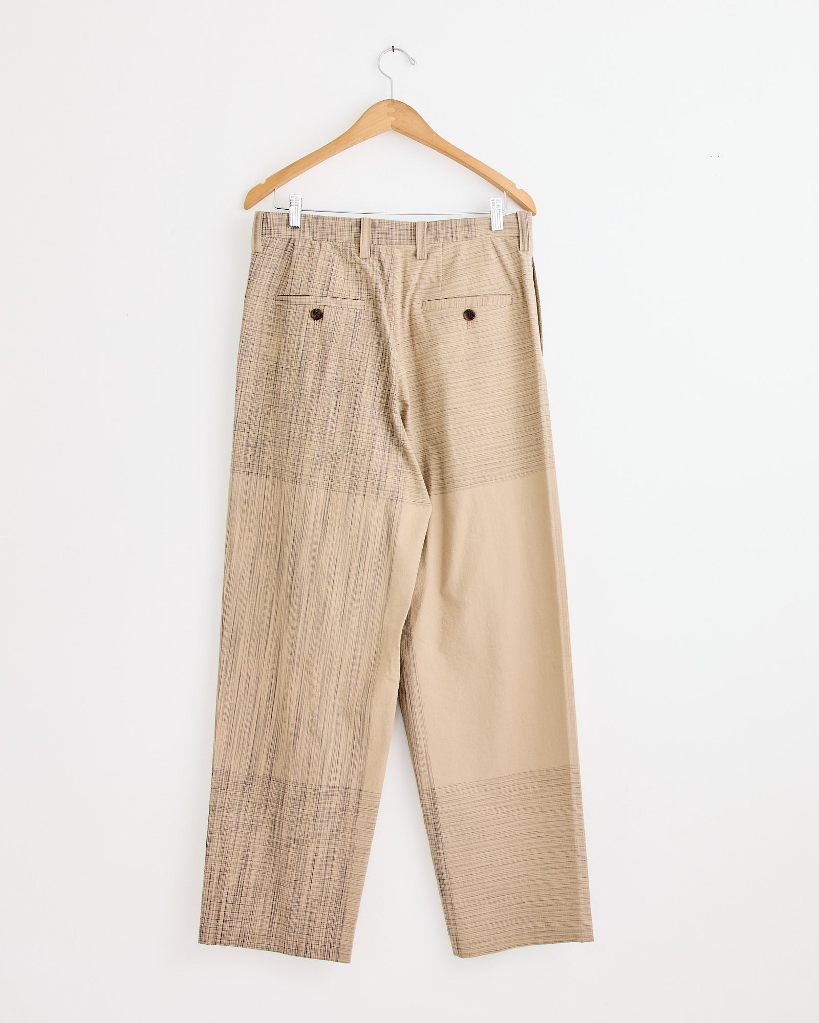 The Kasuri Pant in Beige by IM MEN, featuring solid and plaid panels with buttoned back pockets, hangs on a wooden hanger against a white wall. Chat with us online for any questions about fit or sizing.