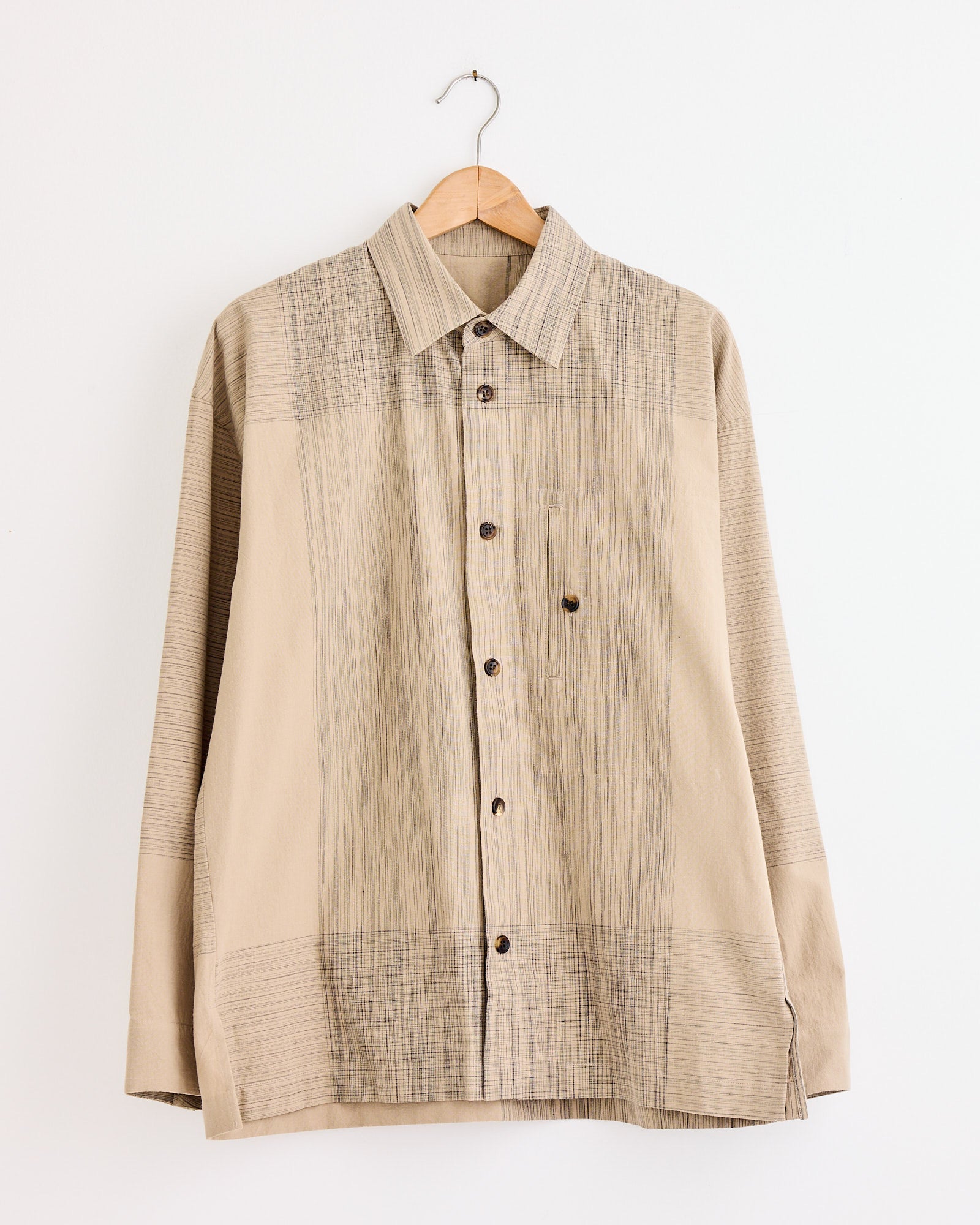The IM MEN Kasuri Shirt in Beige features long sleeves and subtle vertical and horizontal stripes. Displayed on a wooden hanger against a white wall. For fit questions, use our online chat for personalized sizing advice.
