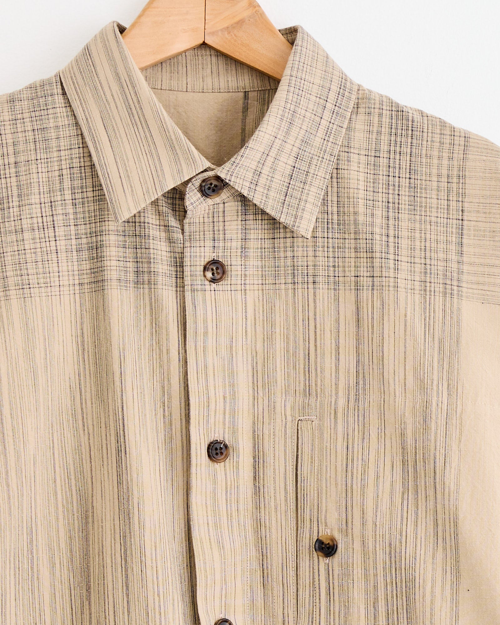 The IM MEN Kasuri Shirt in Beige features thin dark stripes, a chest pocket, and brown buttons. Shown on a wooden hanger against a white background. Chat with us online for sizing help.