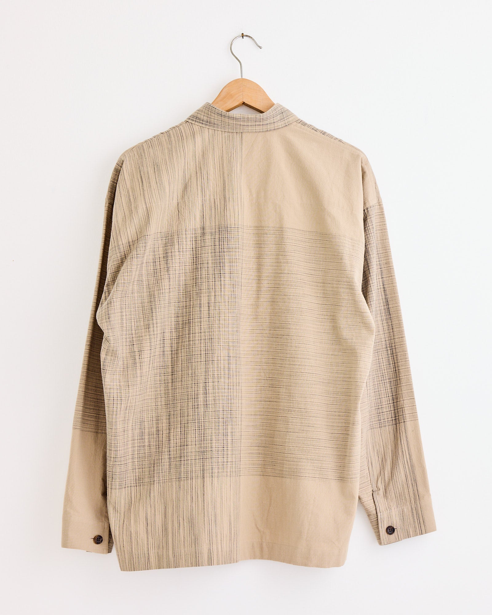 The IM MEN Kasuri Shirt in Beige features thin vertical and horizontal lines and is shown from the back on a wooden hanger. For sizing or fit questions, our online chat is ready to assist you.