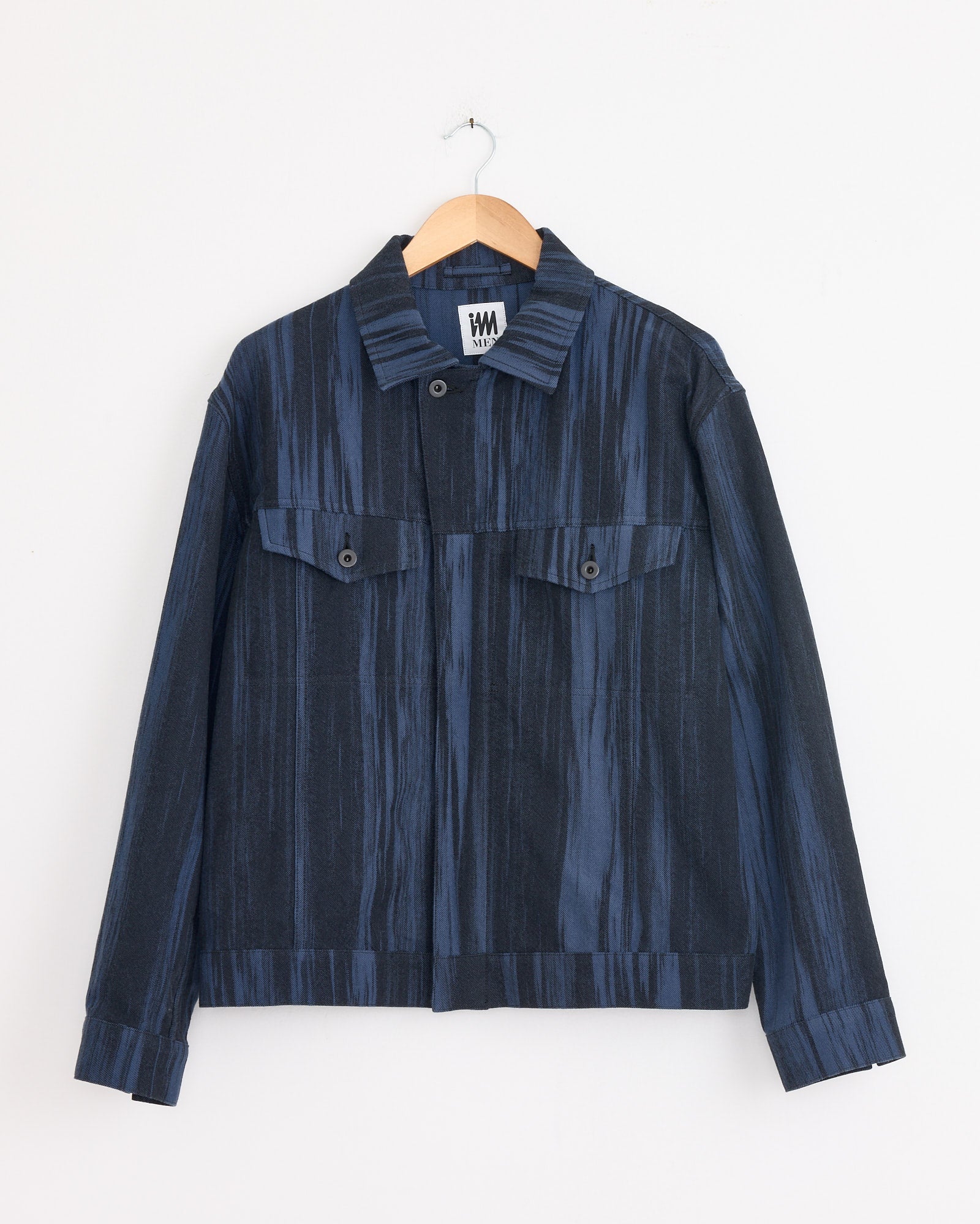 The IM MEN Cotton Jacquard Jacket in Dark Blue features long sleeves, a subtle vertical stripe pattern, buttoned cuffs, flap chest pockets, and a collar. Made in Japan, it's shown on a wooden hanger against a white background.