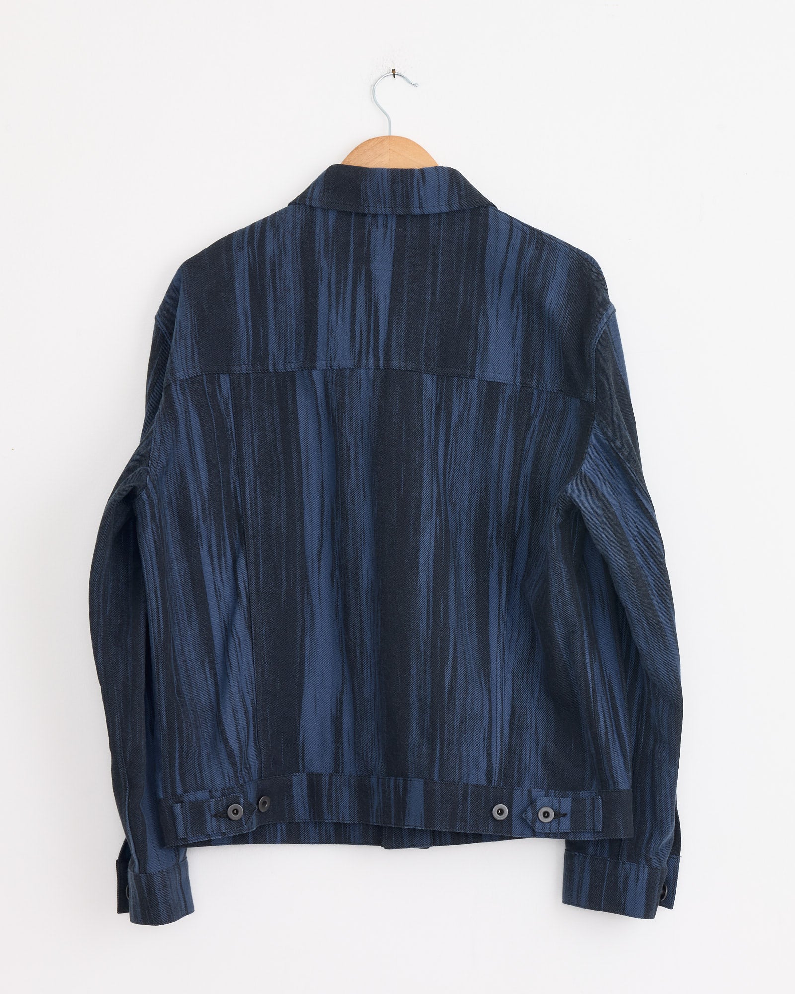The Cotton Jacquard Jacket in Dark Blue by IM MEN hangs on a wooden hanger against a white wall, shown from the back. Made in Japan, it features button details near the waist and a structured collar.