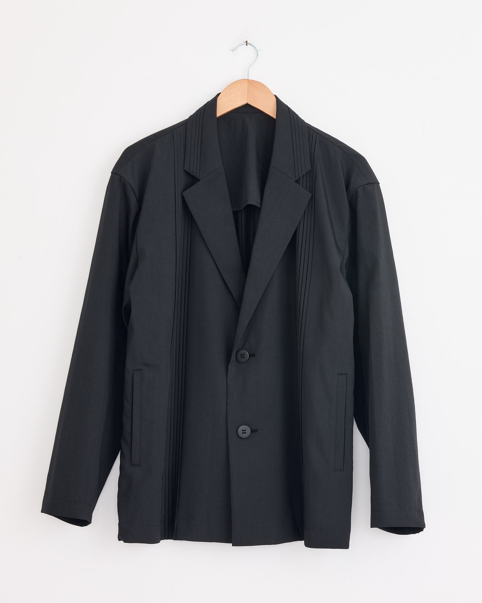 The IM MEN Air Pleats Blazer in Black features a notched lapel, two buttons, and vertical seam details, displayed on a wooden hanger against a plain white wall.