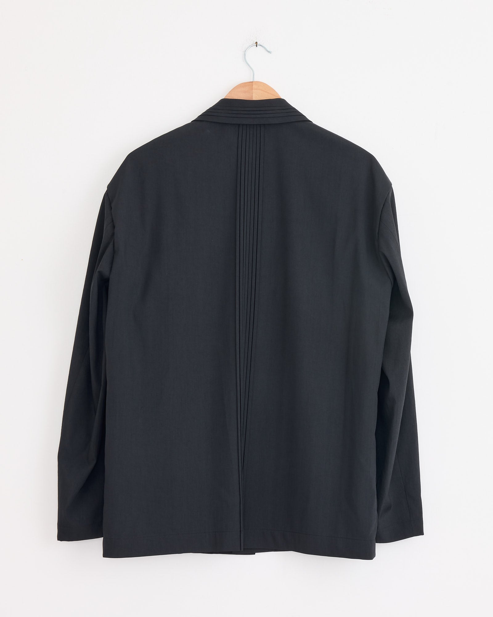 The Air Pleats Blazer in Black by IM MEN hangs on a wooden hanger against a white wall, displayed from the back with vertical center stitching details.