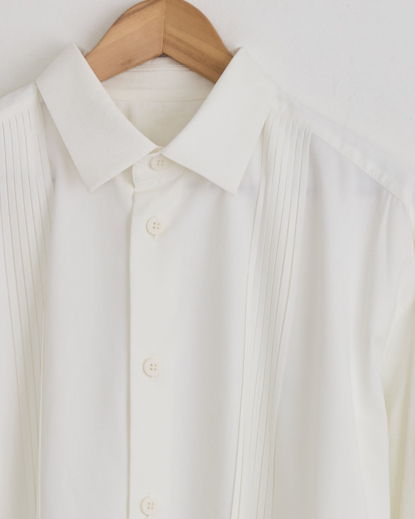 A close-up of the IM MEN Pleats Shirt in White, made in Japan, displayed on a wooden hanger with vertical pleats beside the button placket and a crisp pointed collar.