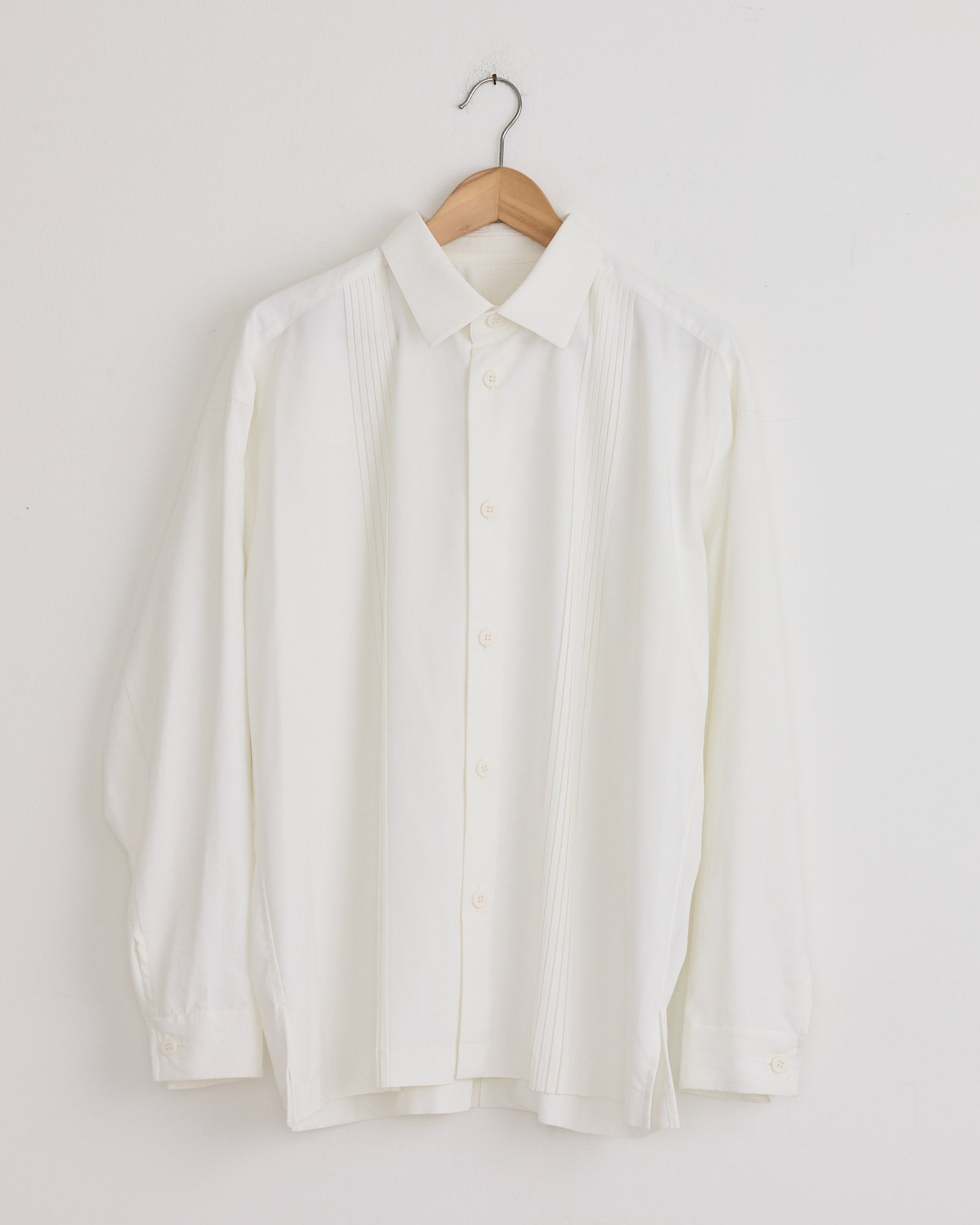 The IM MEN Pleats Shirt in White is a long-sleeved, collared shirt with front vertical pleats, made from recycled polyester, shown on a wooden hanger against a plain white wall.