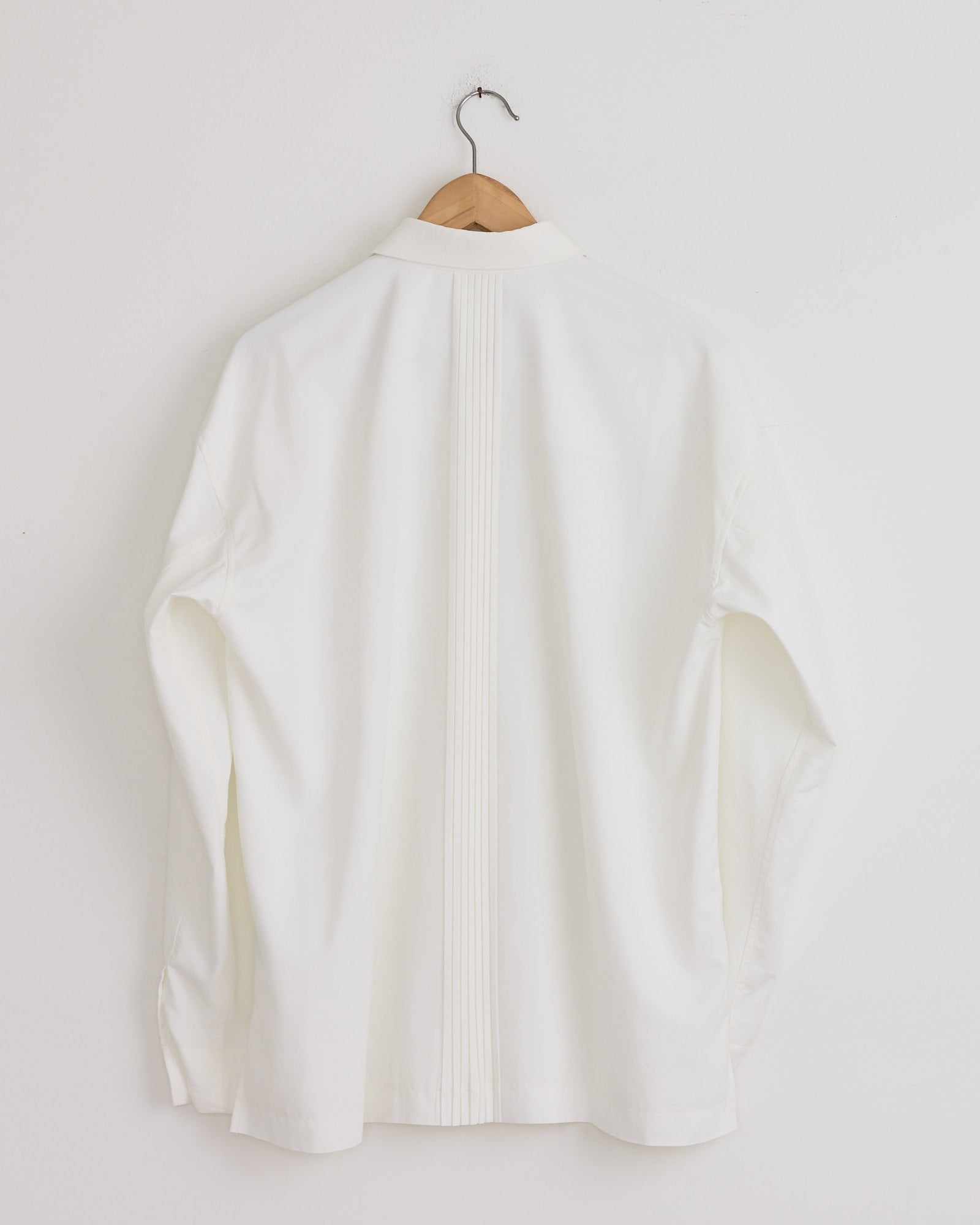 The Pleats Shirt in White by IM MEN hangs on a wooden hanger against a white wall, shown from the back. Made in Japan from recycled polyester, it features vertical center pleats.