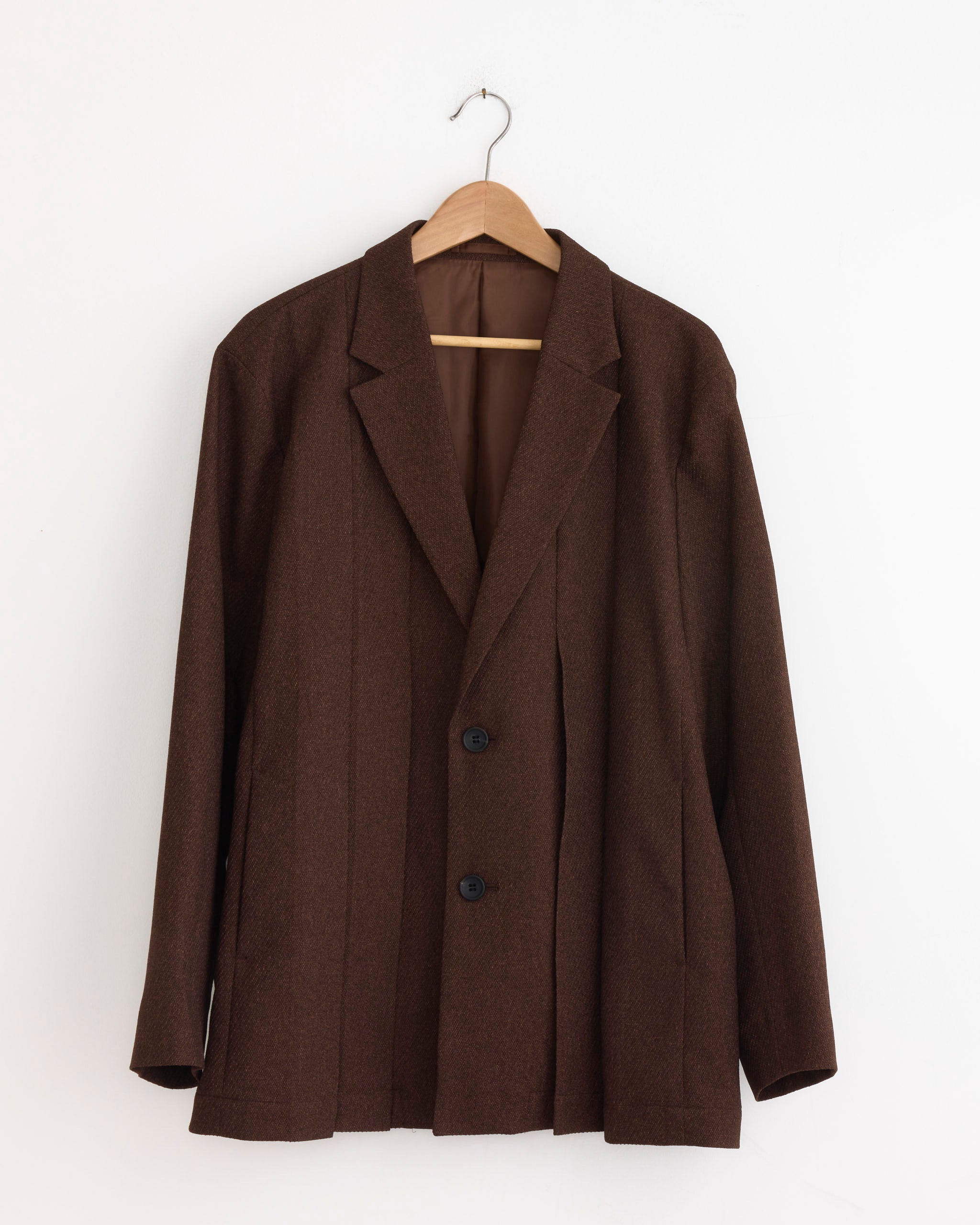 The IM MEN Wool Like Tweed Blazer in Brown, featuring notched lapels and two black buttons, hangs on a wooden hanger against a white wall for a timeless men's look.