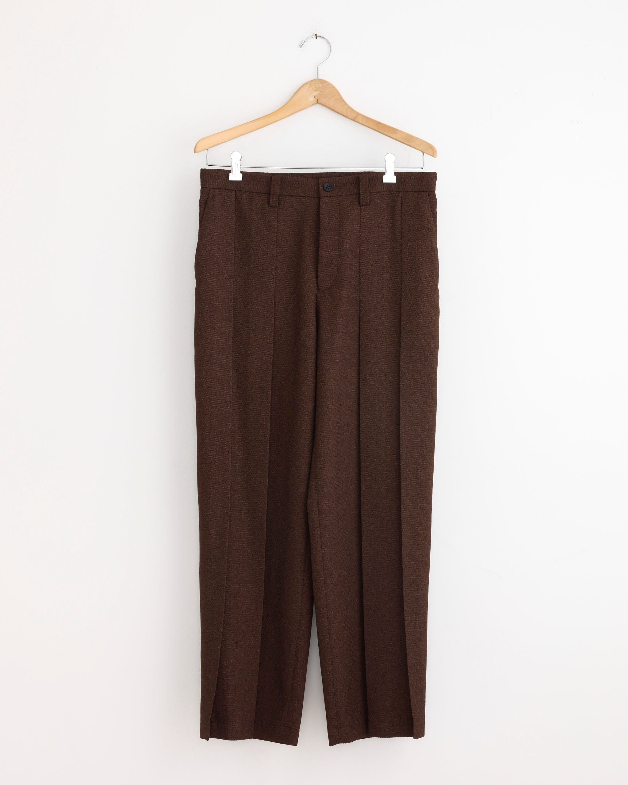 The IM MEN Wool Like Tweed Pant in Brown, featuring a tapered silhouette, hangs on a wooden hanger against a plain white wall.