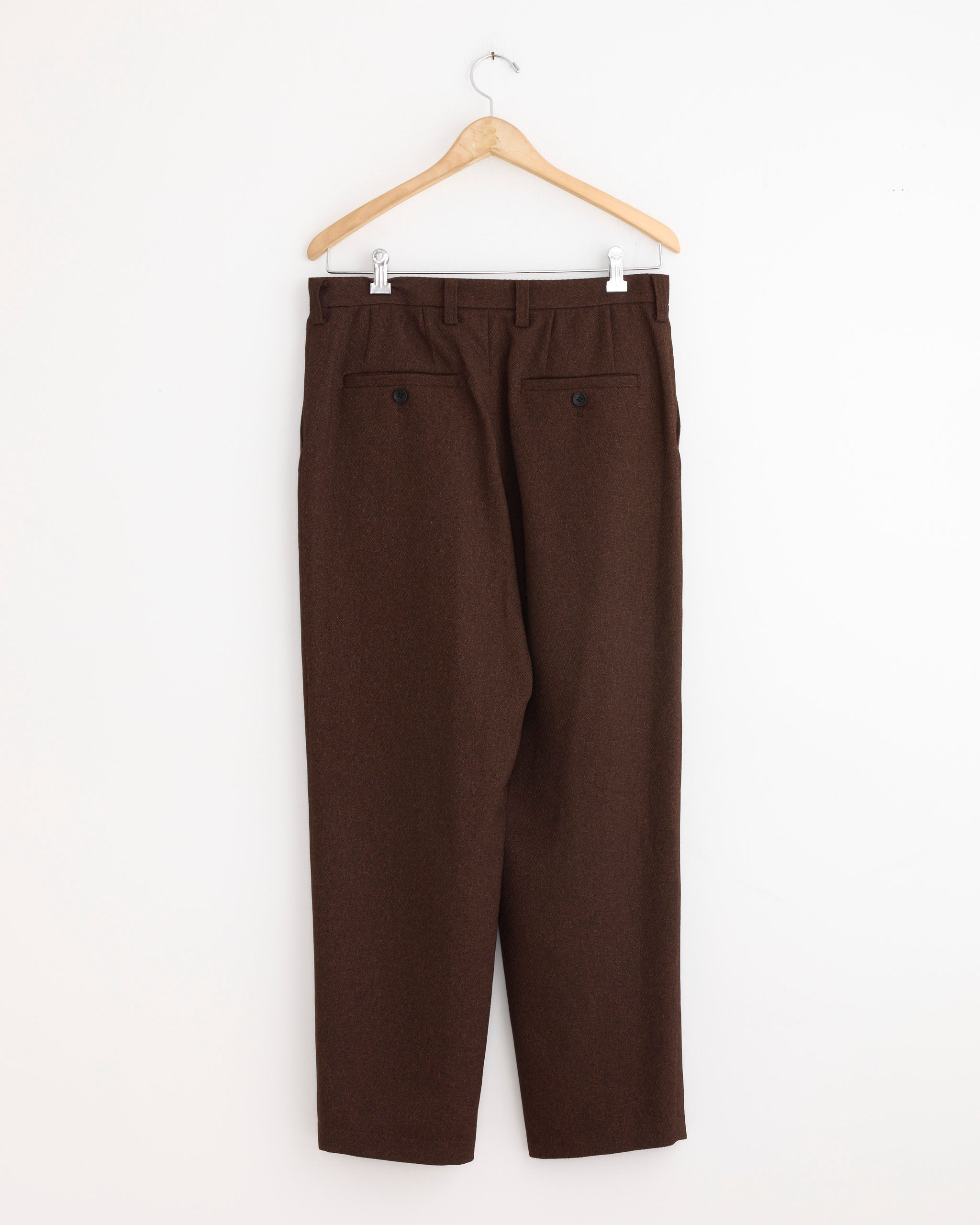 The IM MEN Wool Like Tweed Pant in Brown, featuring a tapered silhouette and two buttoned back pockets, is displayed hanging on a wooden hanger against a white wall, shown from the back.