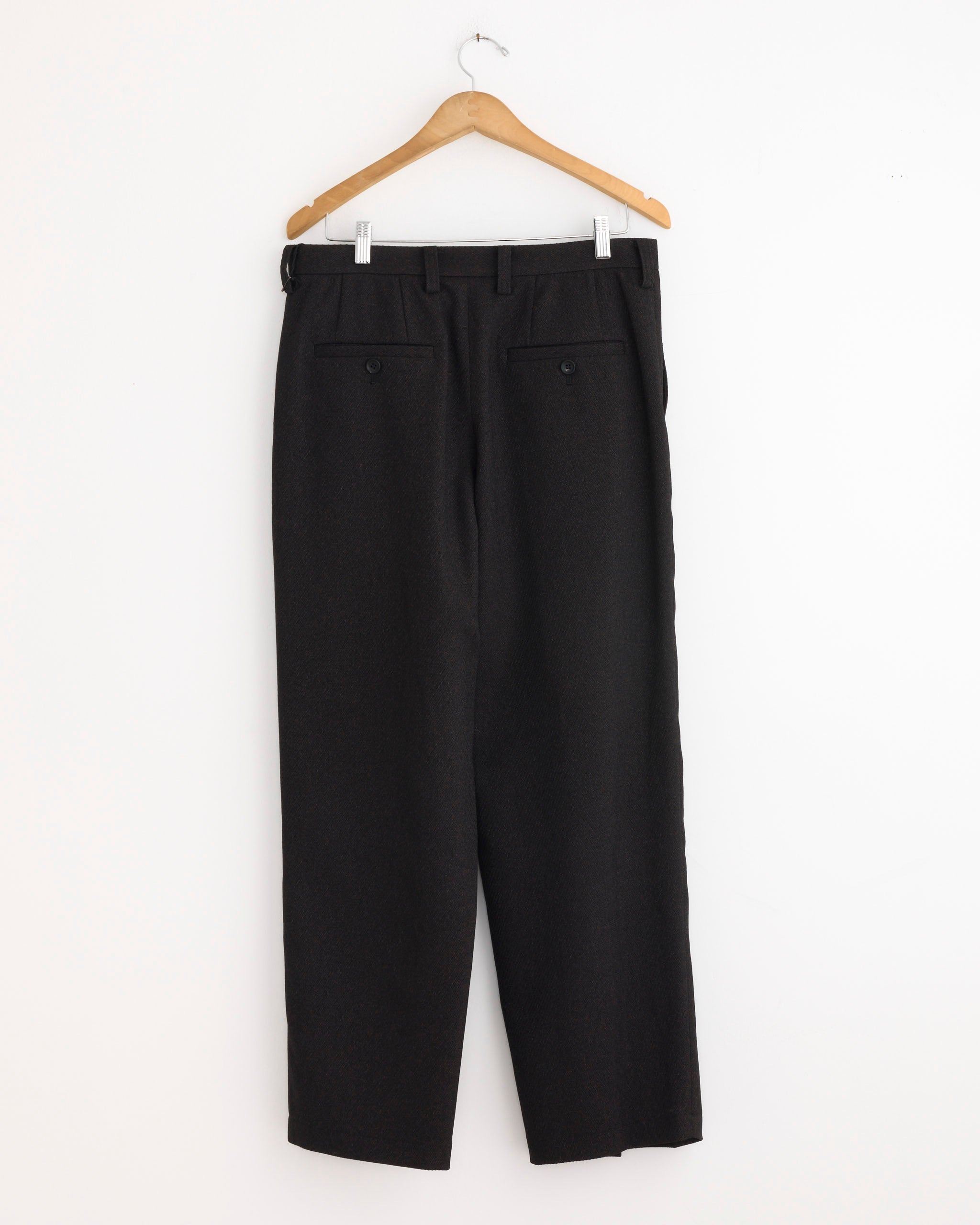 A pair of IM MEN Wool Like Tweed Pant in Dark Brown hangs on a wooden hanger against a plain white wall, shown from the back with two buttoned pockets and a refined tapered silhouette.