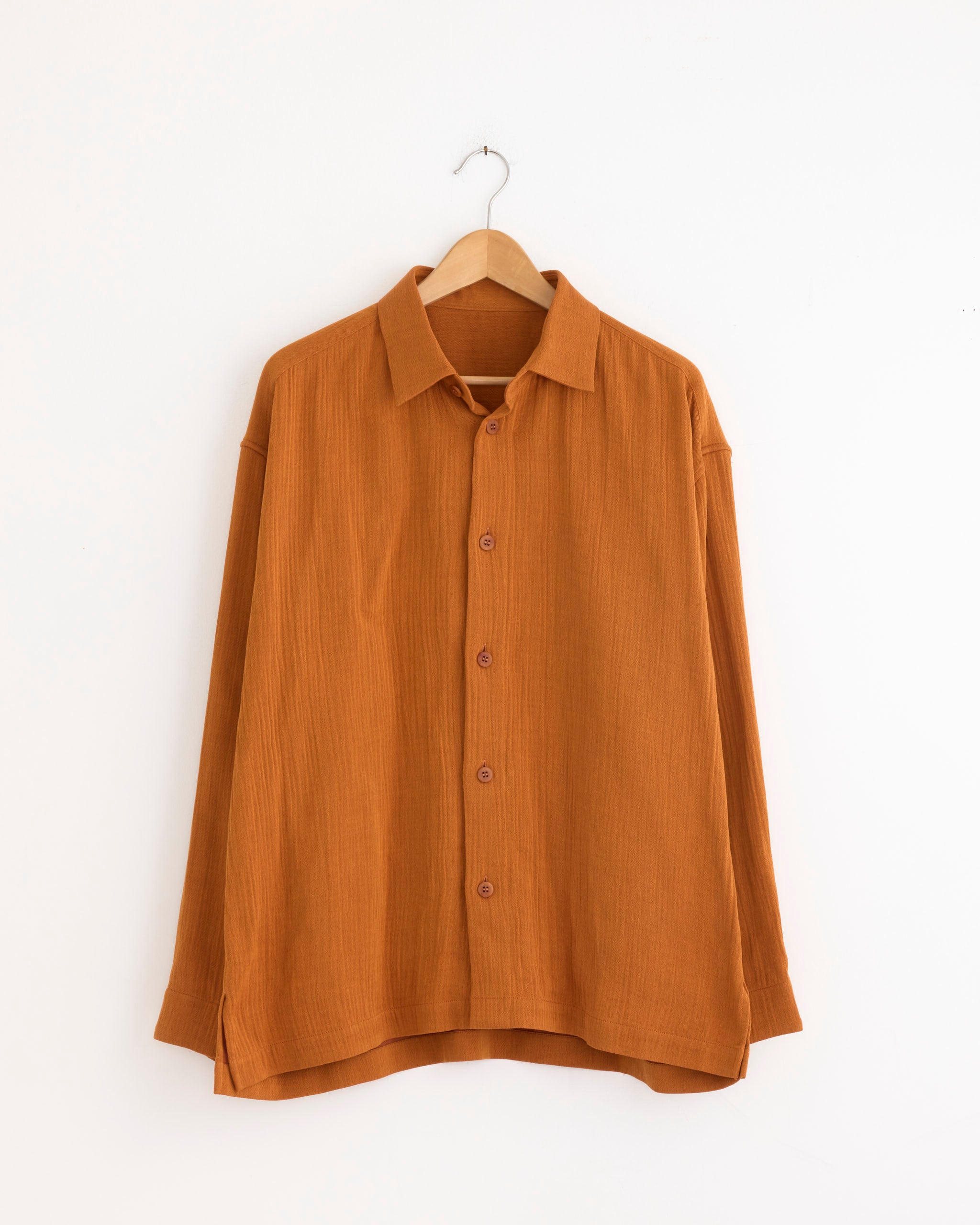 The IM MEN Double Weave Crepe Shirt in Dark Orange features long sleeves, button-up front, and lightweight textured fabric. Displayed on a wooden hanger with a white background. For sizing questions, contact us via online chat.