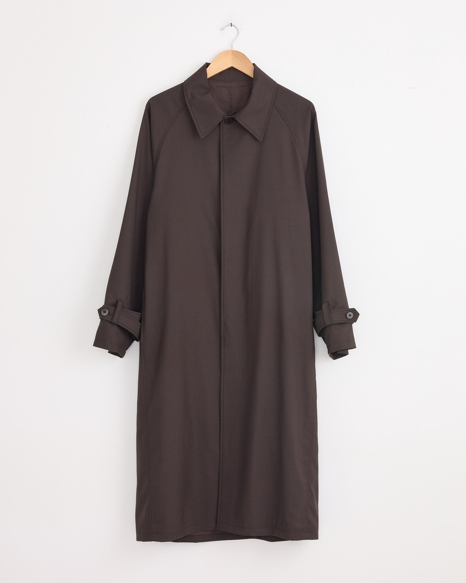 The Stoffa Raglan Coat in Chocolate, crafted from luxurious merino wool in Italy, with a collar and buttoned cuffs, hangs on a wooden hanger against a plain white wall.