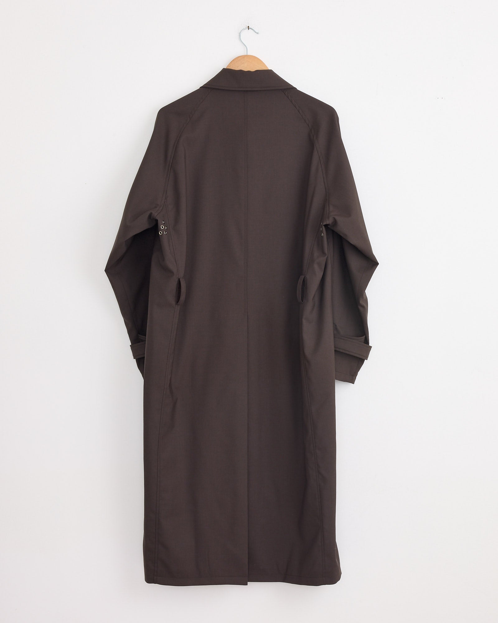 The Stoffa Raglan Coat in Chocolate, made in Italy, hangs on a wooden hanger against a white wall, displayed from the back with slightly folded sleeves.