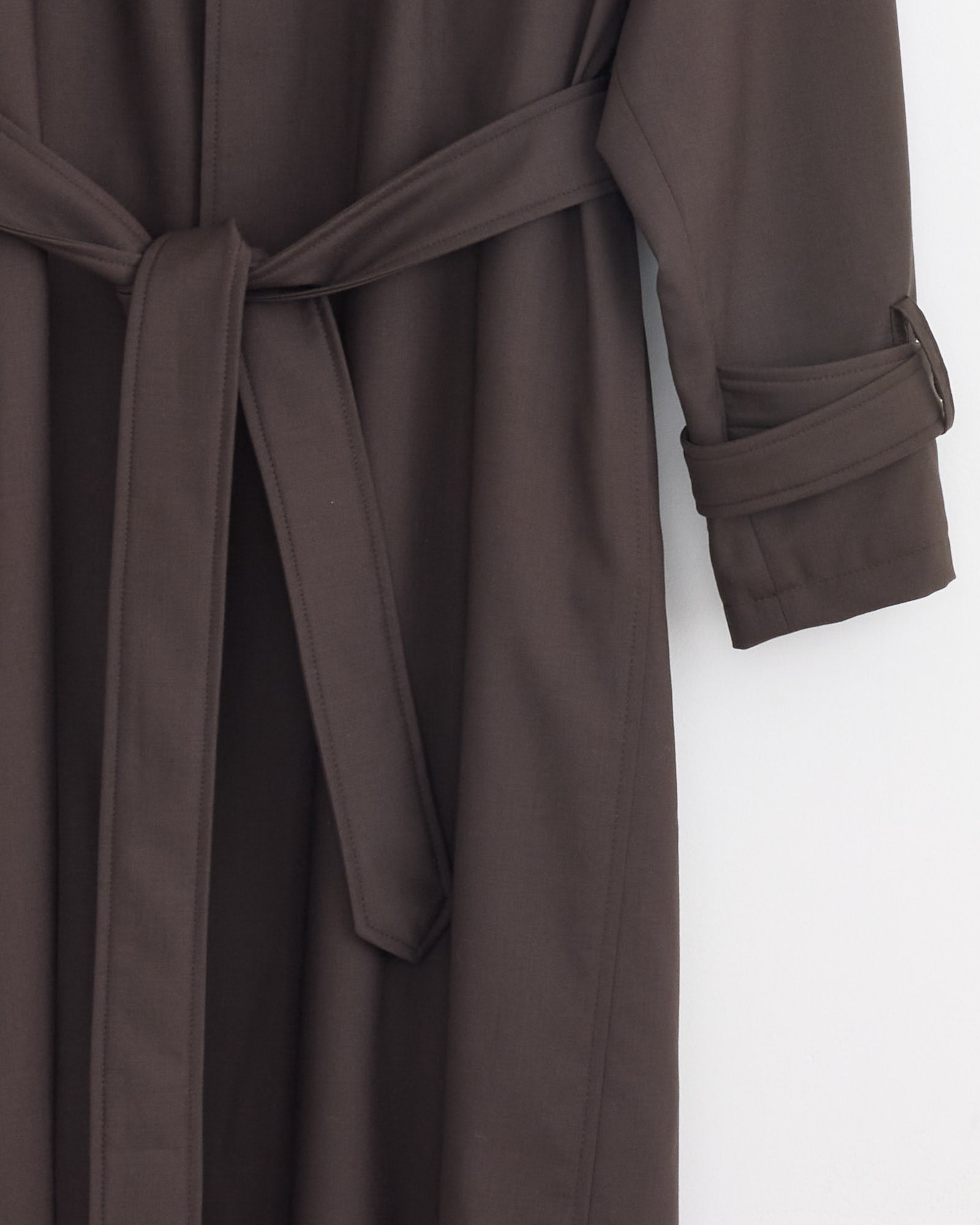 A close-up of the Stoffa Raglan Coat in Chocolate, made from merino wool, features a fabric belt tied at the waist and a sleeve with a buttoned strap detail, set against a light background.