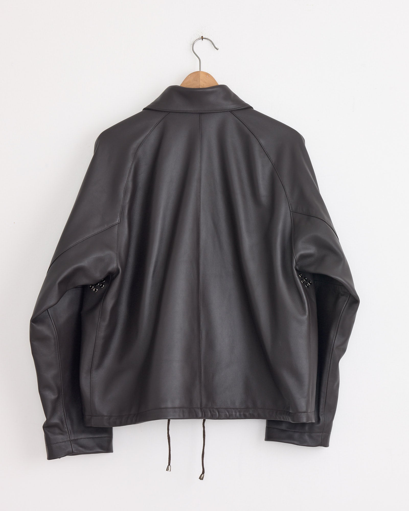 The Stoffa Field Blouson in Chocolate, crafted in Italy, hangs on a wooden hanger against a white wall, shown from the back with gently bent sleeves and visible bottom hem drawstrings to highlight its refined craftsmanship.