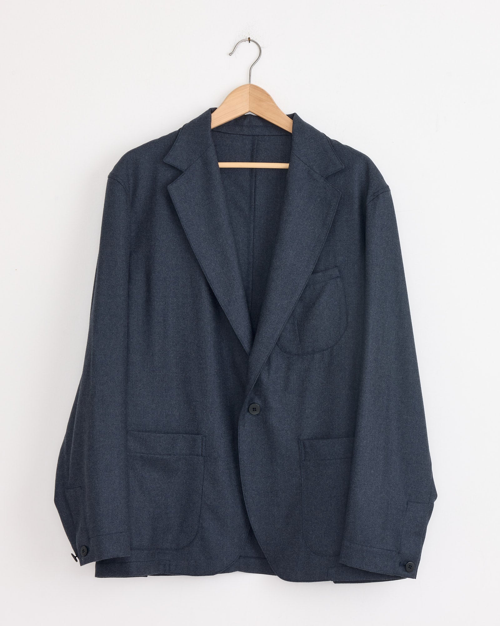 The Stoffa Single Breasted Shirt Jacket in Indigo, featuring three front pockets, is displayed on a wooden hanger against a white wall.