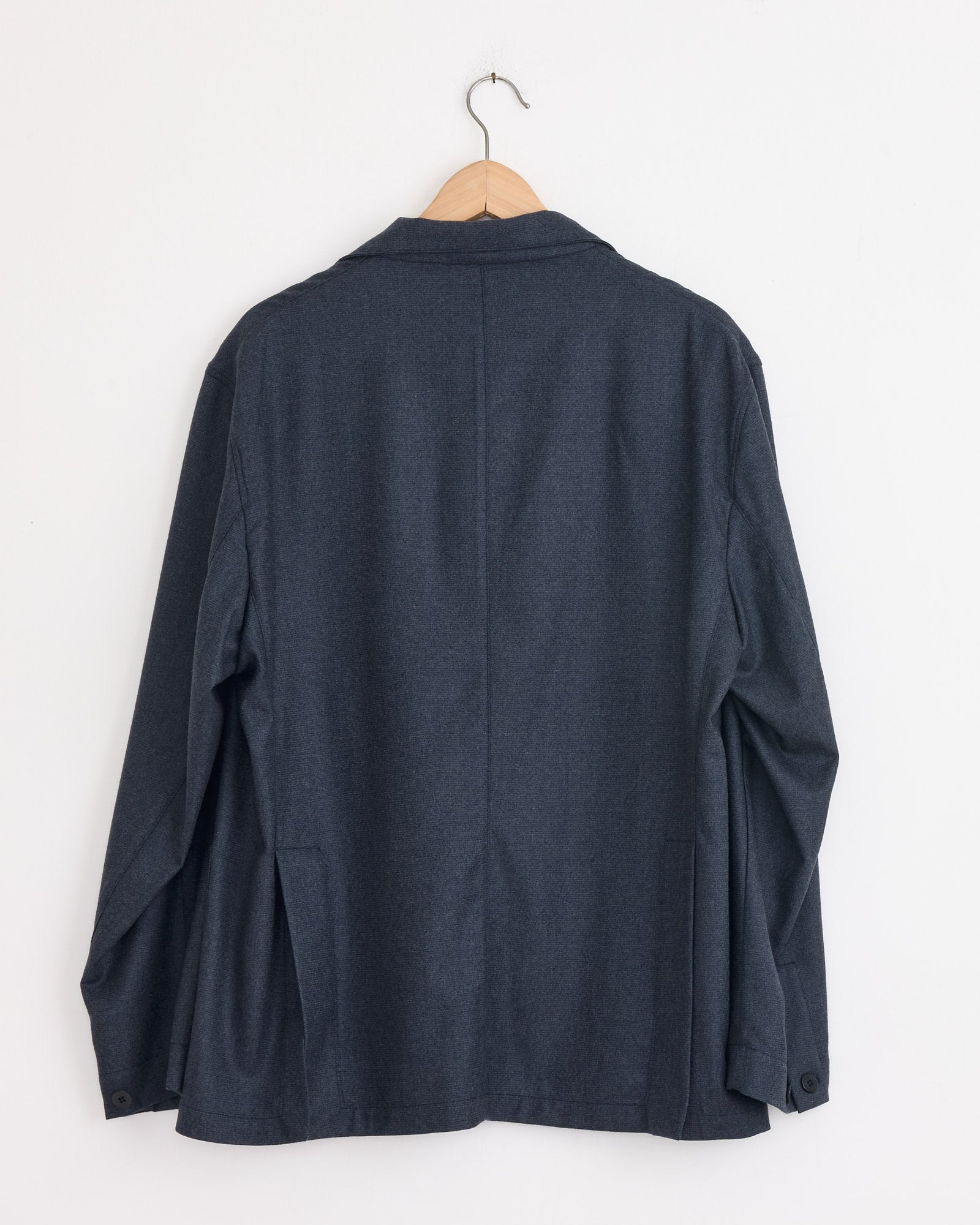 The Stoffa Single Breasted Shirt Jacket in Indigo, made in Italy, hangs on a wooden hanger against a plain white wall, displayed from the back.