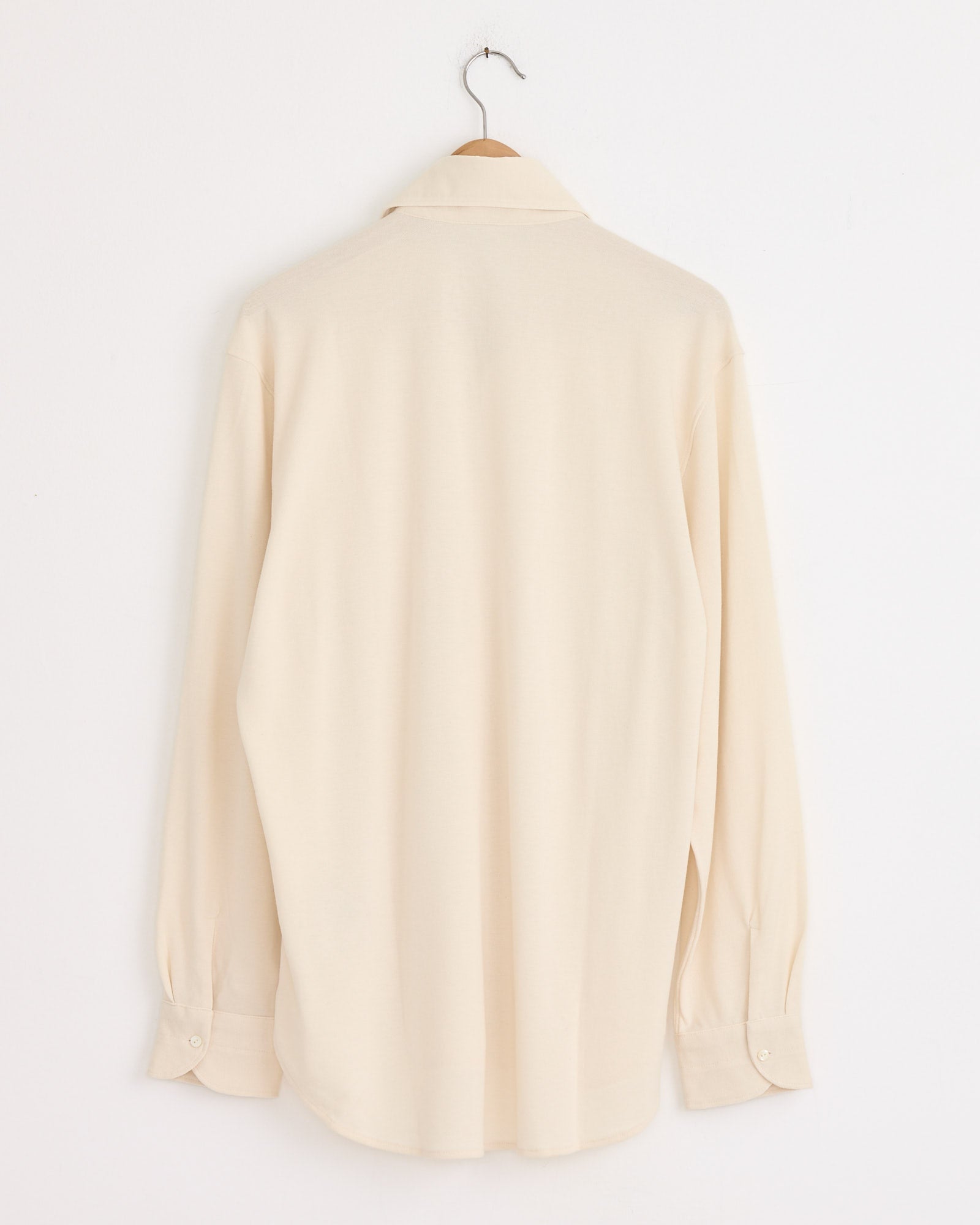 The Stoffa Spread Collar Shirt in Ivory, crafted from a cotton-silk blend with long sleeves, hangs from a wooden hanger against a plain white wall, displayed from the back.