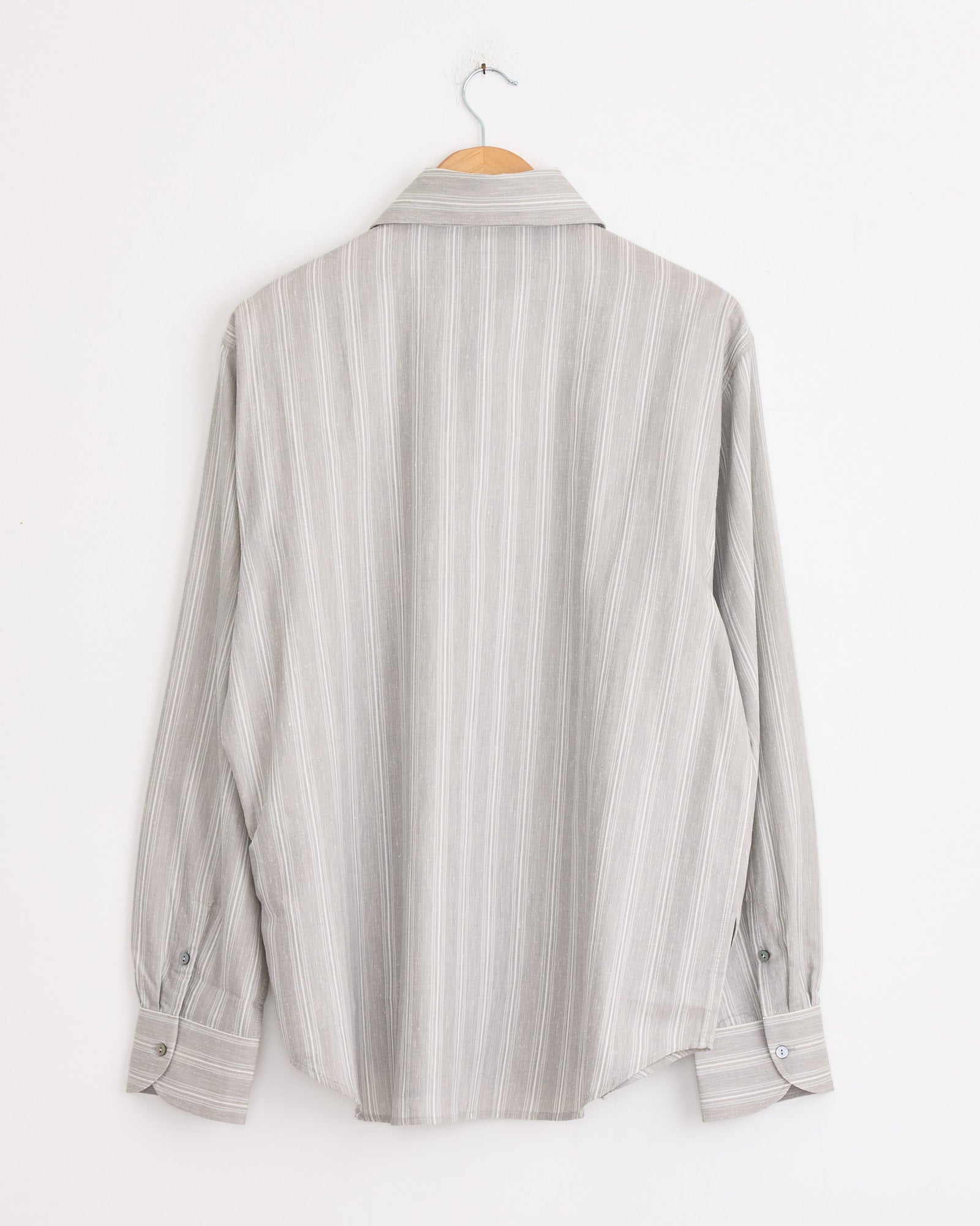 The Stoffa Spread Collar Shirt in Steel Grey/Pearl Stripe, made in Italy, hangs on a wooden hanger against a white wall. Shown from the back, it features long sleeves and vertical white stripes.