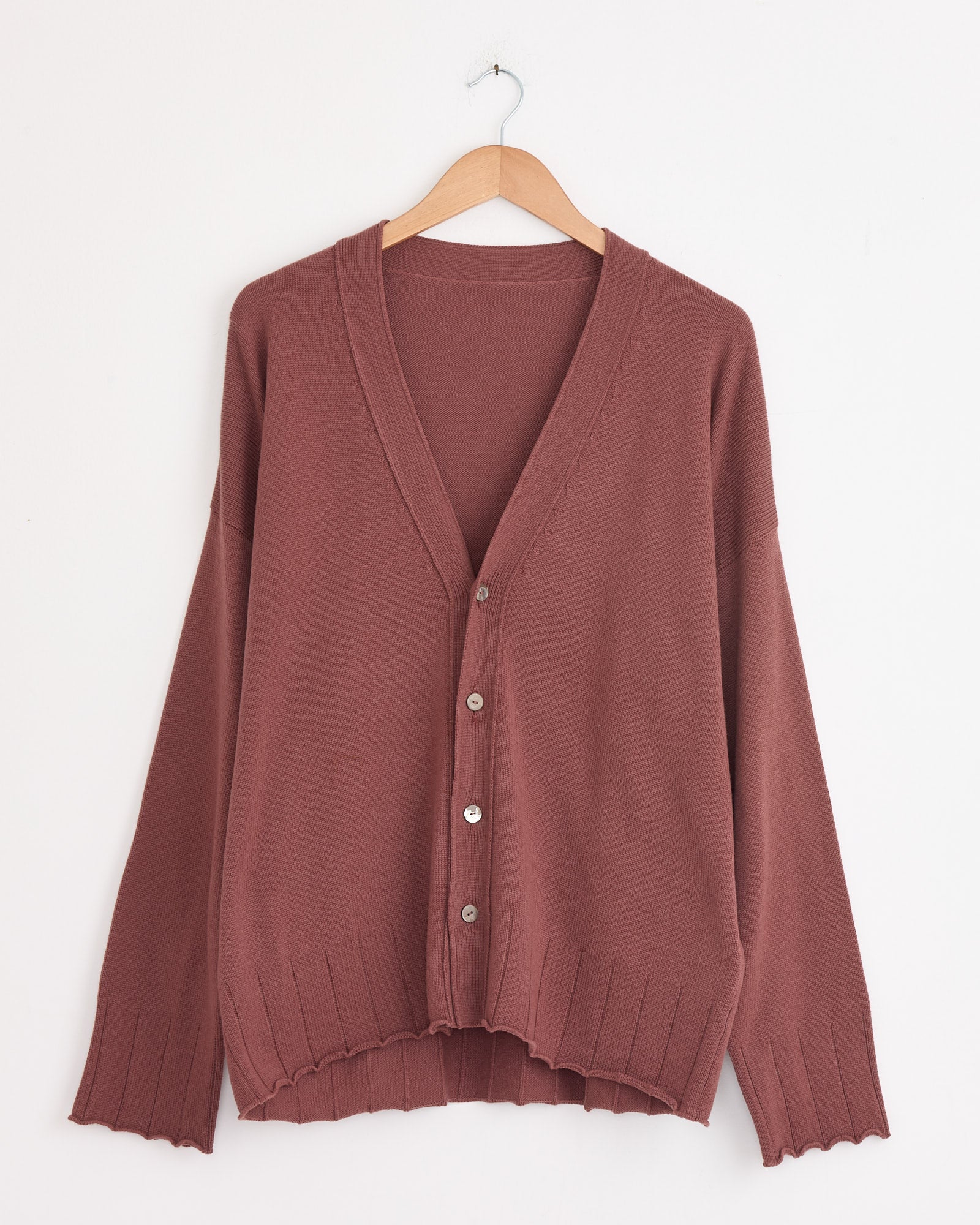 A brick-colored, 100% cashmere Stoffa Drop Shoulder Cardigan with a V-neckline hangs on a wooden hanger against a white wall. Made in Italy, it features ribbed cuffs and hem, and four silver buttons down the front.