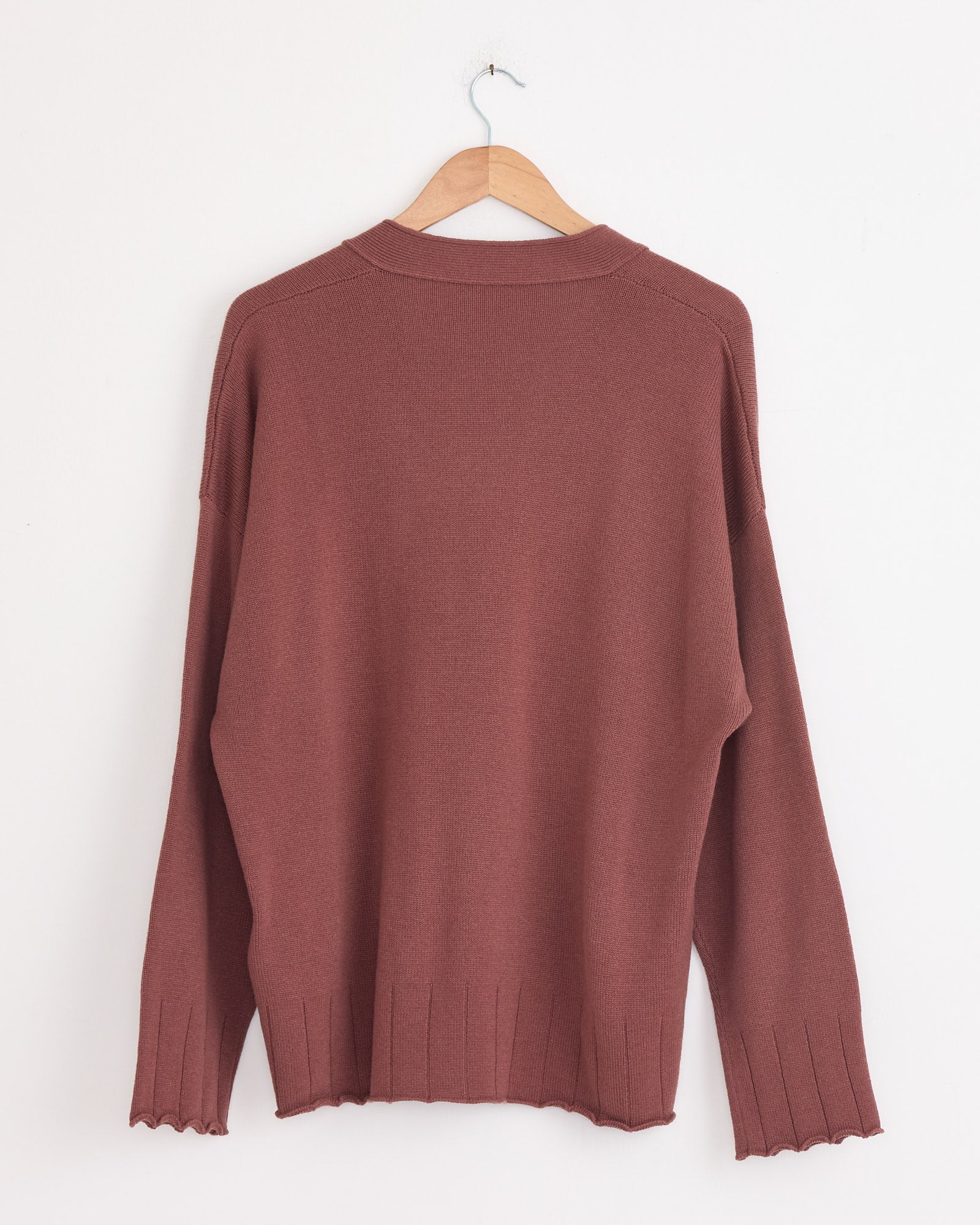 The Stoffa Drop Shoulder Cardigan in Brick, made in Italy from 100% cashmere, features long sleeves, ribbed cuffs and hem, a relaxed fit, and minimal detailing. Shown hanging on a wooden hanger against a white wall.