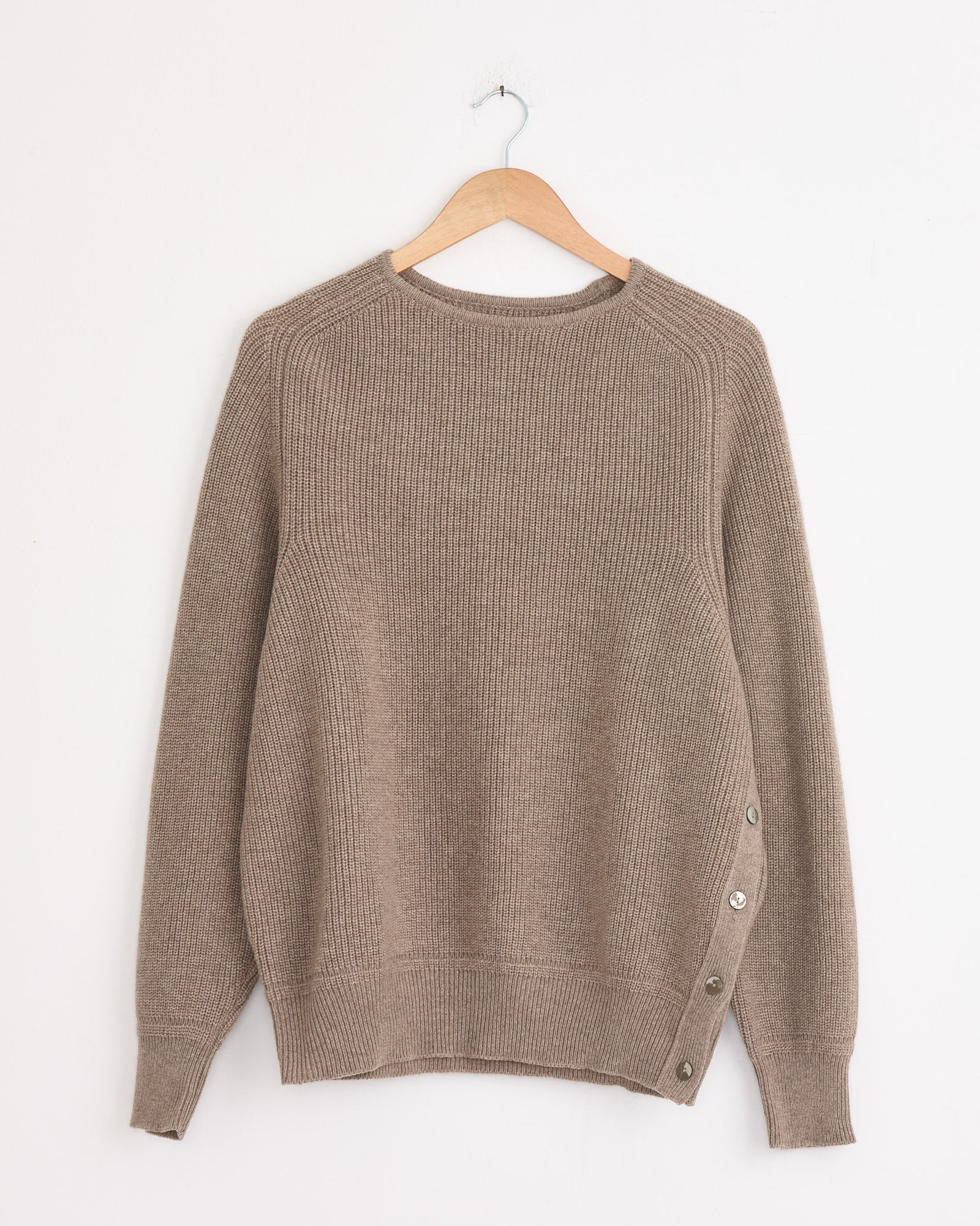 A Stoffa Side Button Crewneck in Undyed Taupe hangs on a wooden hanger. This cashmere sweater features long sleeves, a ribbed texture, crew neckline, and decorative side buttons near one hem.
