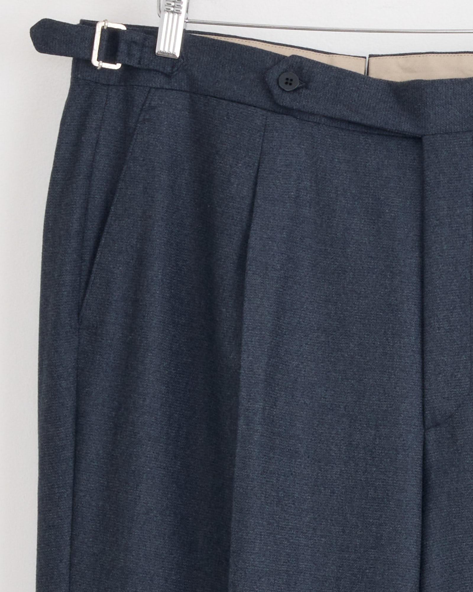 Close-up of Stoffa Single Pleat Trouser in Indigo hanging on a hanger, featuring a waistband with side buckle, button closure, side pocket details, and light inner waistband lining. Made in Italy.