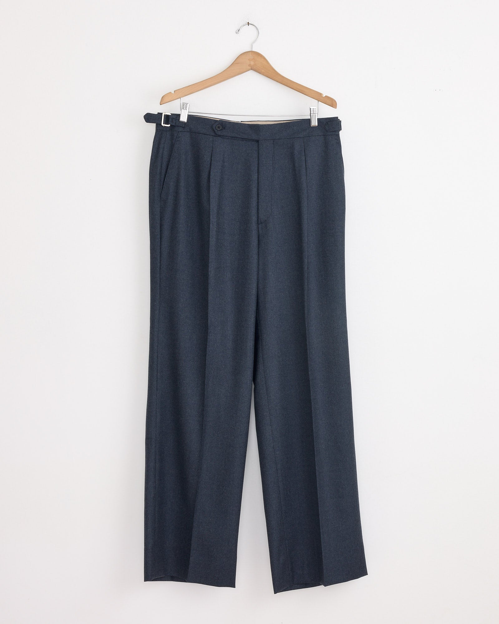 The Stoffa Single Pleat Trouser in Indigo, crafted in Italy, hangs on a wooden hanger against a plain white wall.