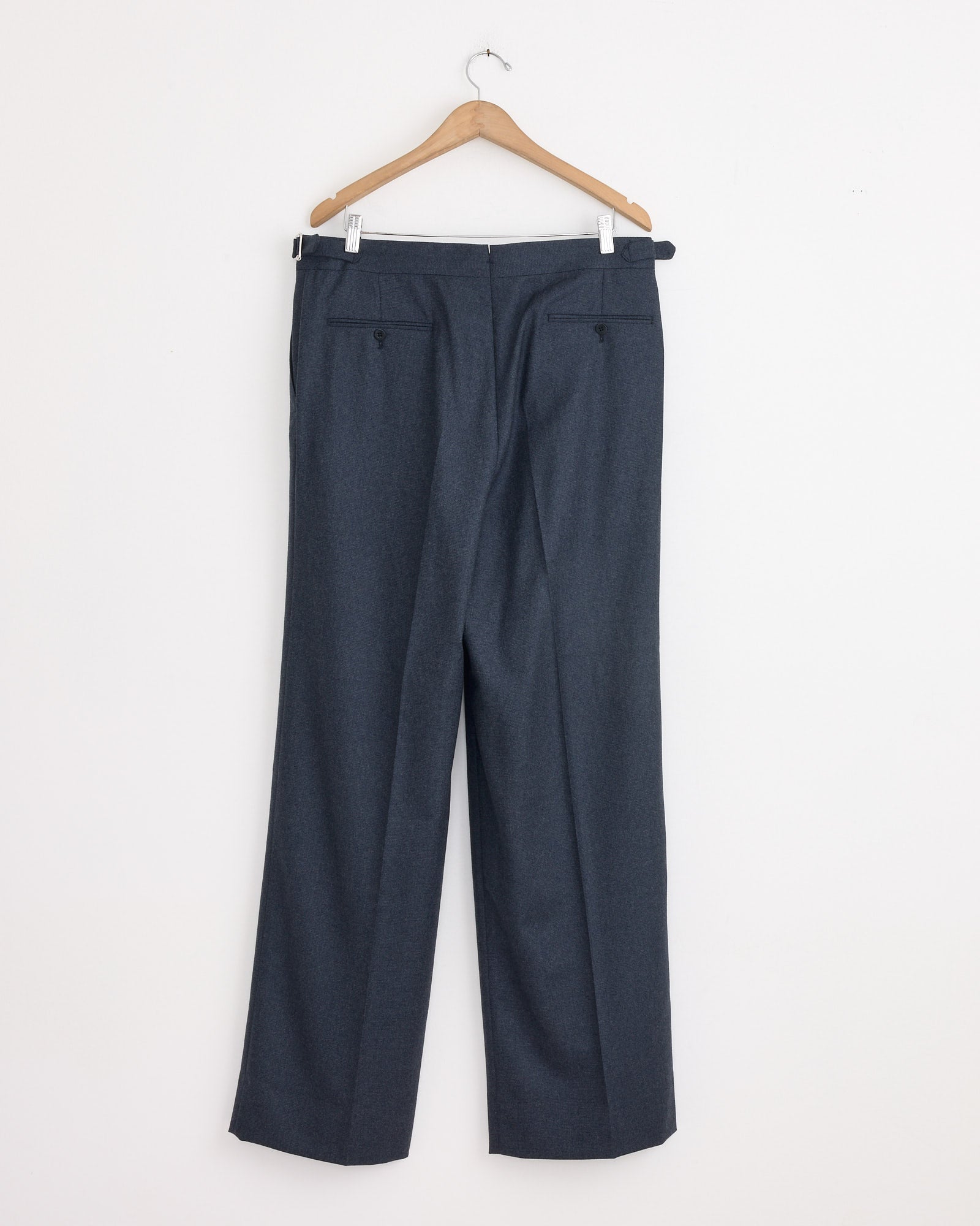 The Stoffa Single Pleat Trouser in Indigo hangs from a wooden hanger against a plain white wall, showcasing the back side with visible belt loops and back pockets.