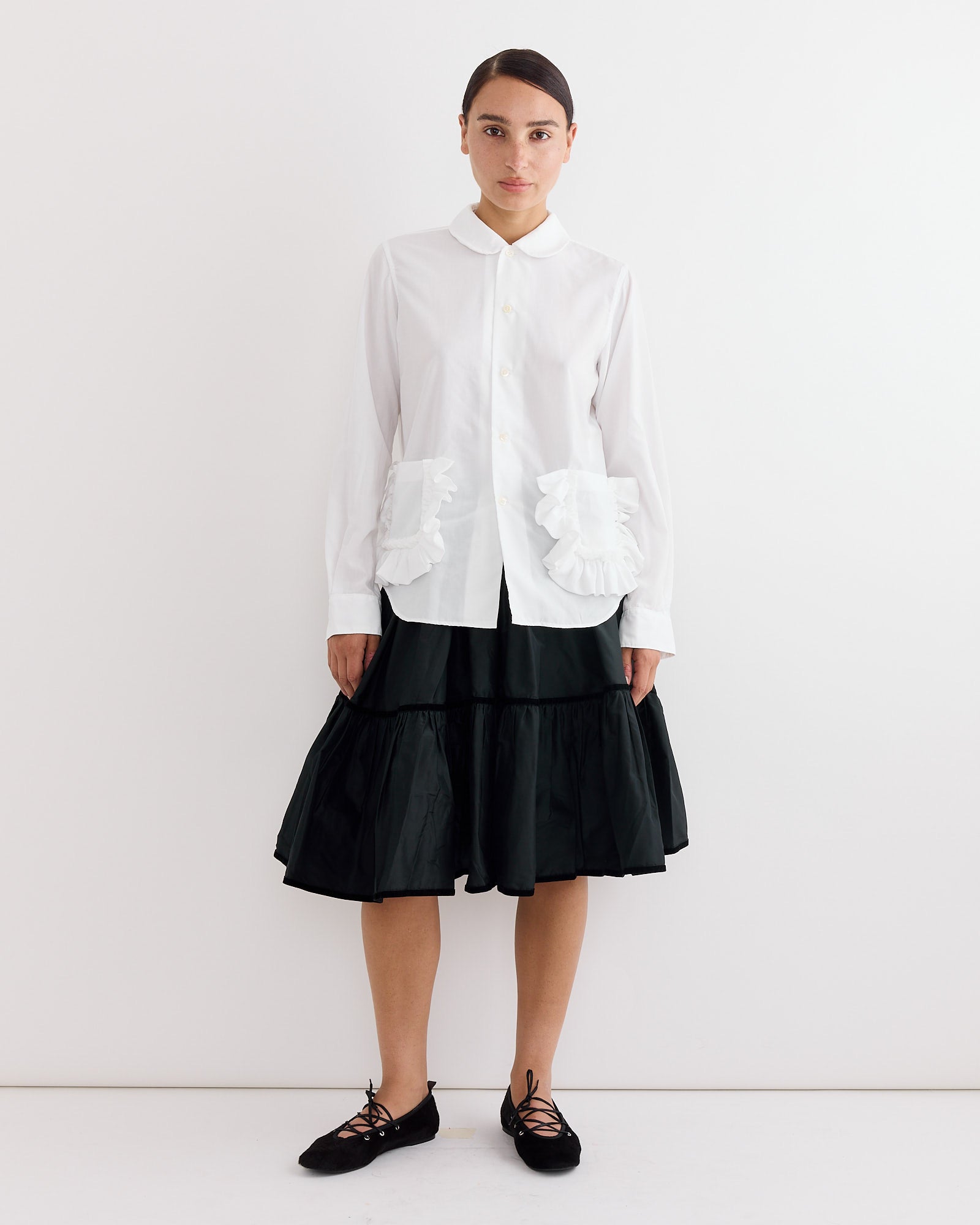 A woman with dark hair tied back wears the Comme Des Garçons Girl Frilled Pocket Blouse in White against a plain background, paired with a black knee-length tiered skirt and black lace-up shoes.