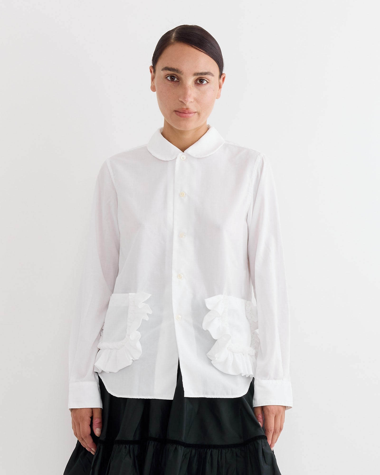 A person with dark hair wears the Comme Des Garçons Girl Frilled Pocket Blouse in White and a black skirt, standing against a plain white background.
