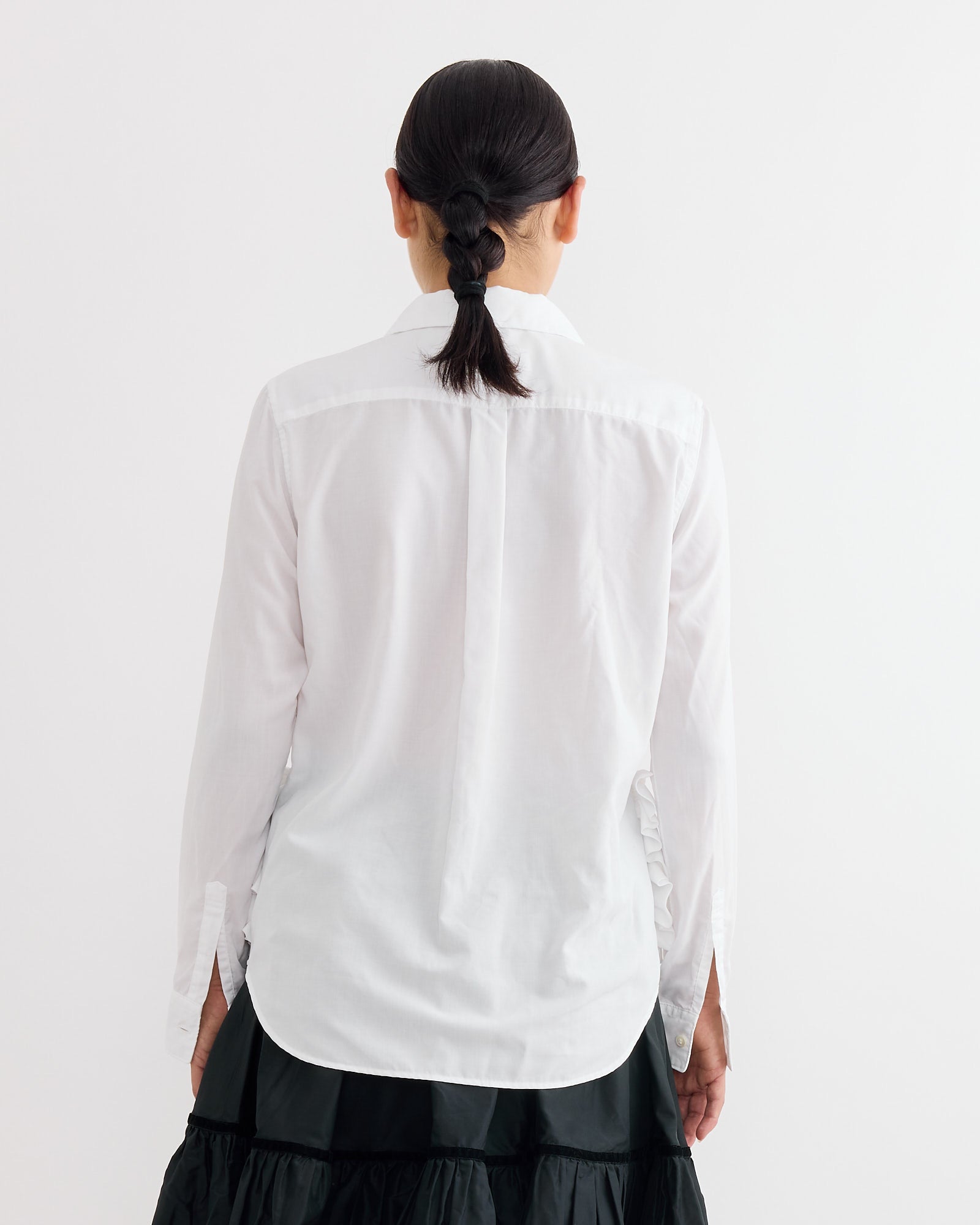 A person with dark hair in a low braid is shown from behind, wearing the Comme Des Garçons Girl Frilled Pocket Blouse in White and a black skirt against a plain white background.