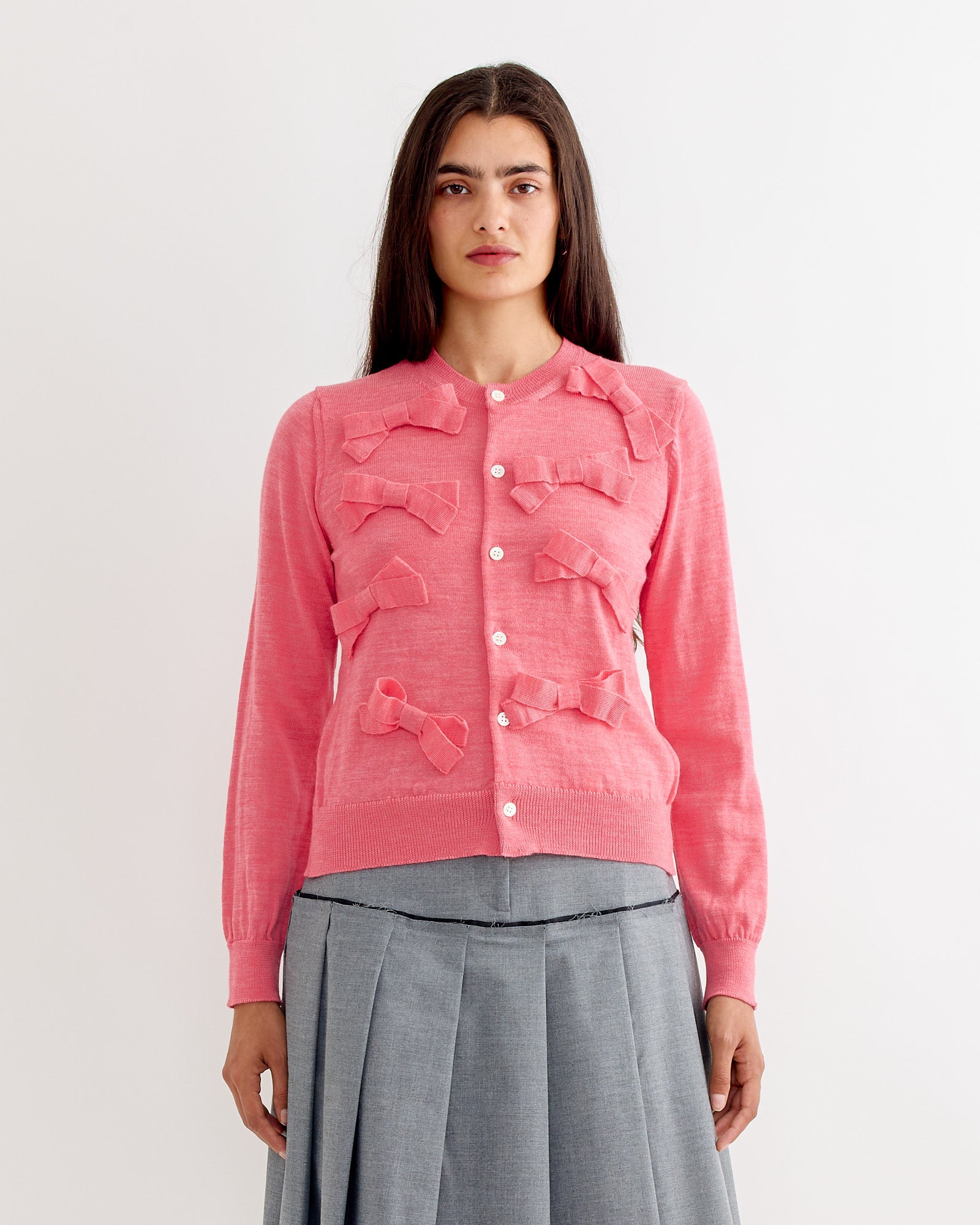 A woman with long dark hair wears the Comme Des Garçons Girl Bow Cardigan in Pink, featuring fabric bows and white buttons, paired with a gray pleated skirt, standing against a plain white background.
