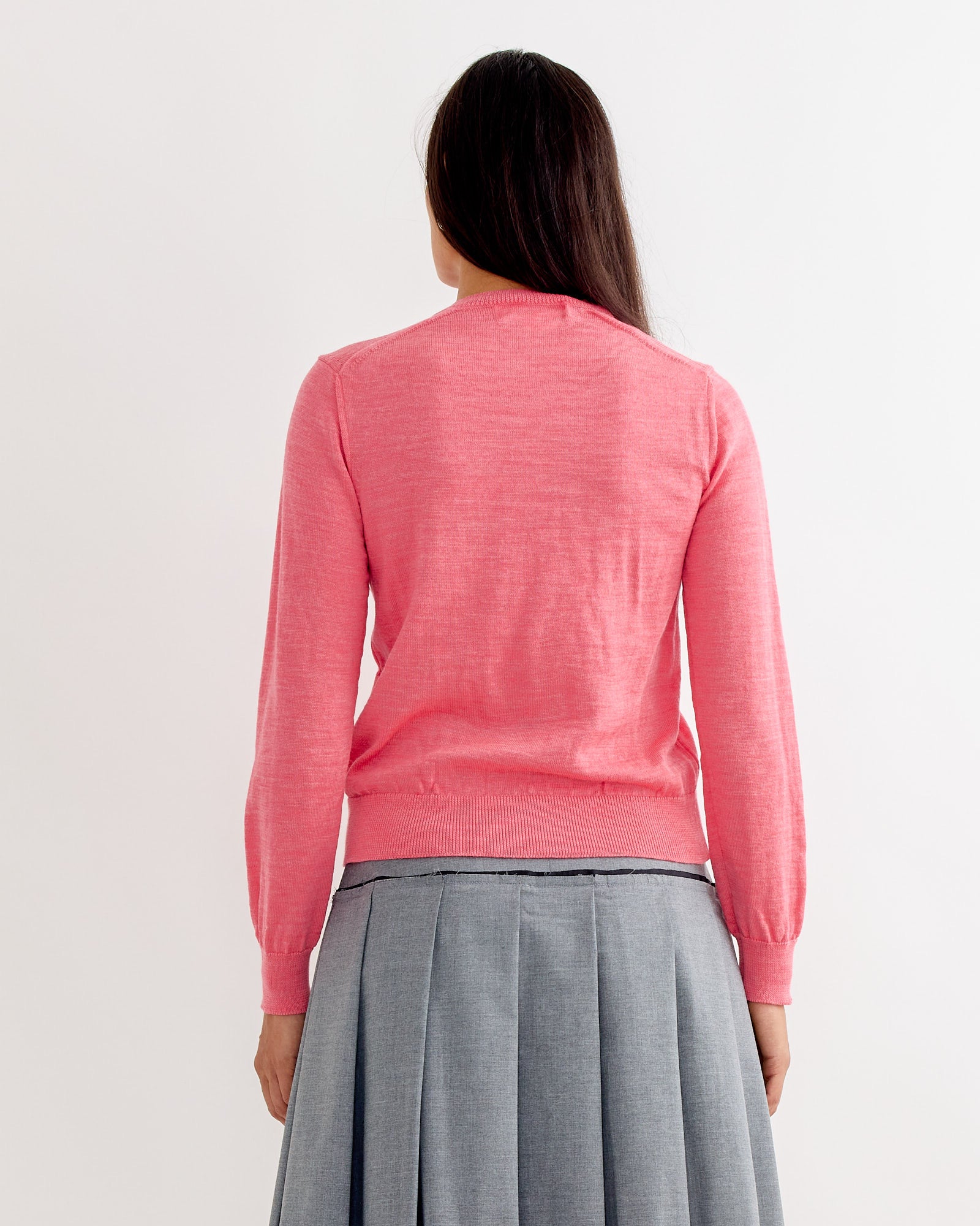 A person with long dark hair wears the Bow Cardigan in Pink by Comme Des Garçons Girl and a gray pleated skirt, standing with their back to the camera against a plain light background.