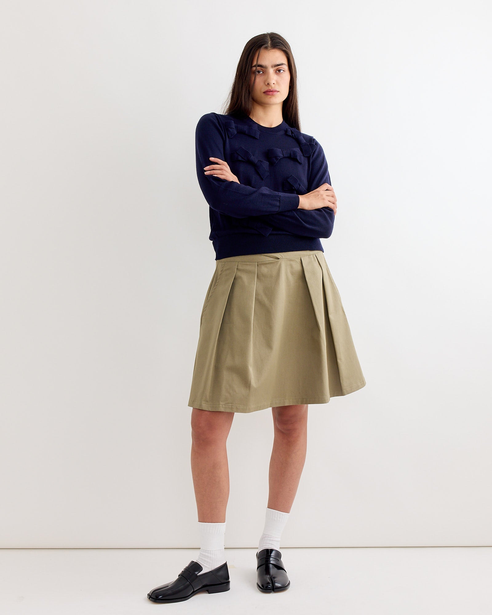 A person with long dark hair stands with arms crossed, wearing the Comme Des Garçons Girl Bow Sweater in Navy, a khaki pleated skirt, white socks, and black loafers against a plain white background.