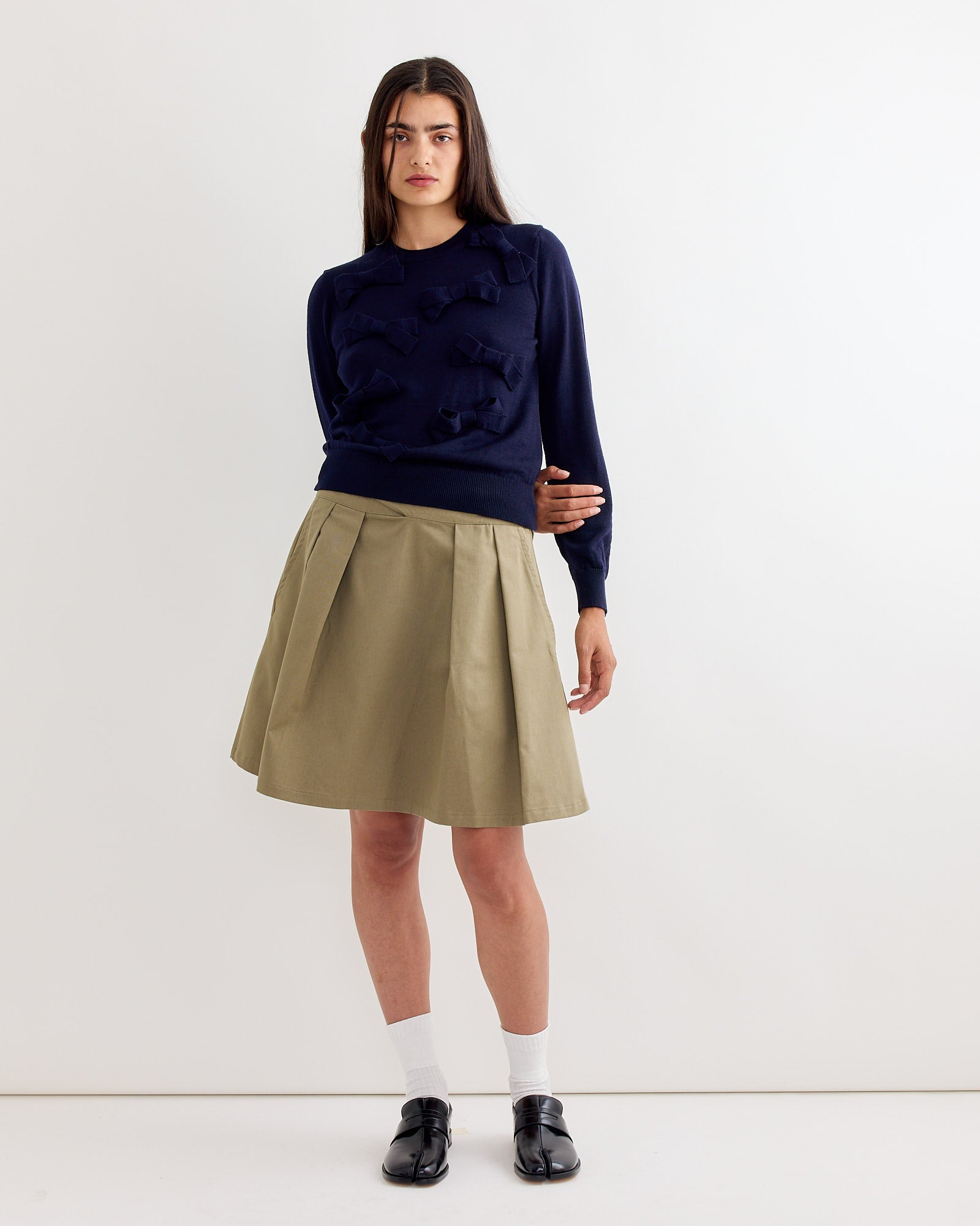 A woman with long straight hair and a neutral expression wears a navy blue sweater with bow details, white socks, black loafers, and the Gimaguas Martin Midi Skirt in Light Khaki while standing against a plain white wall.
