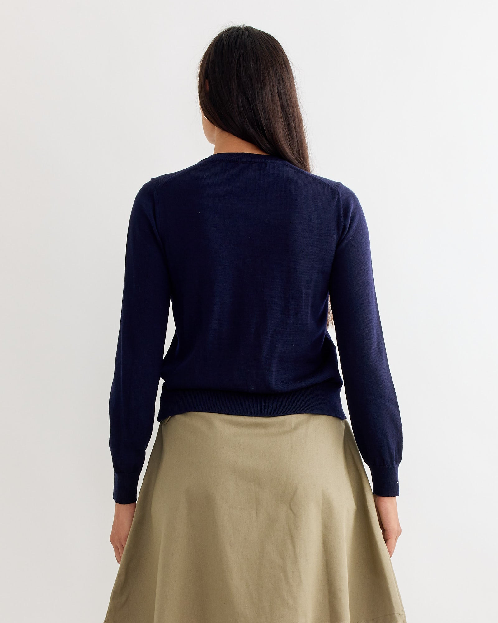 A person with long dark hair, seen from behind against a white background, wears the Comme Des Garçons Girl Bow Sweater in navy with a beige skirt.