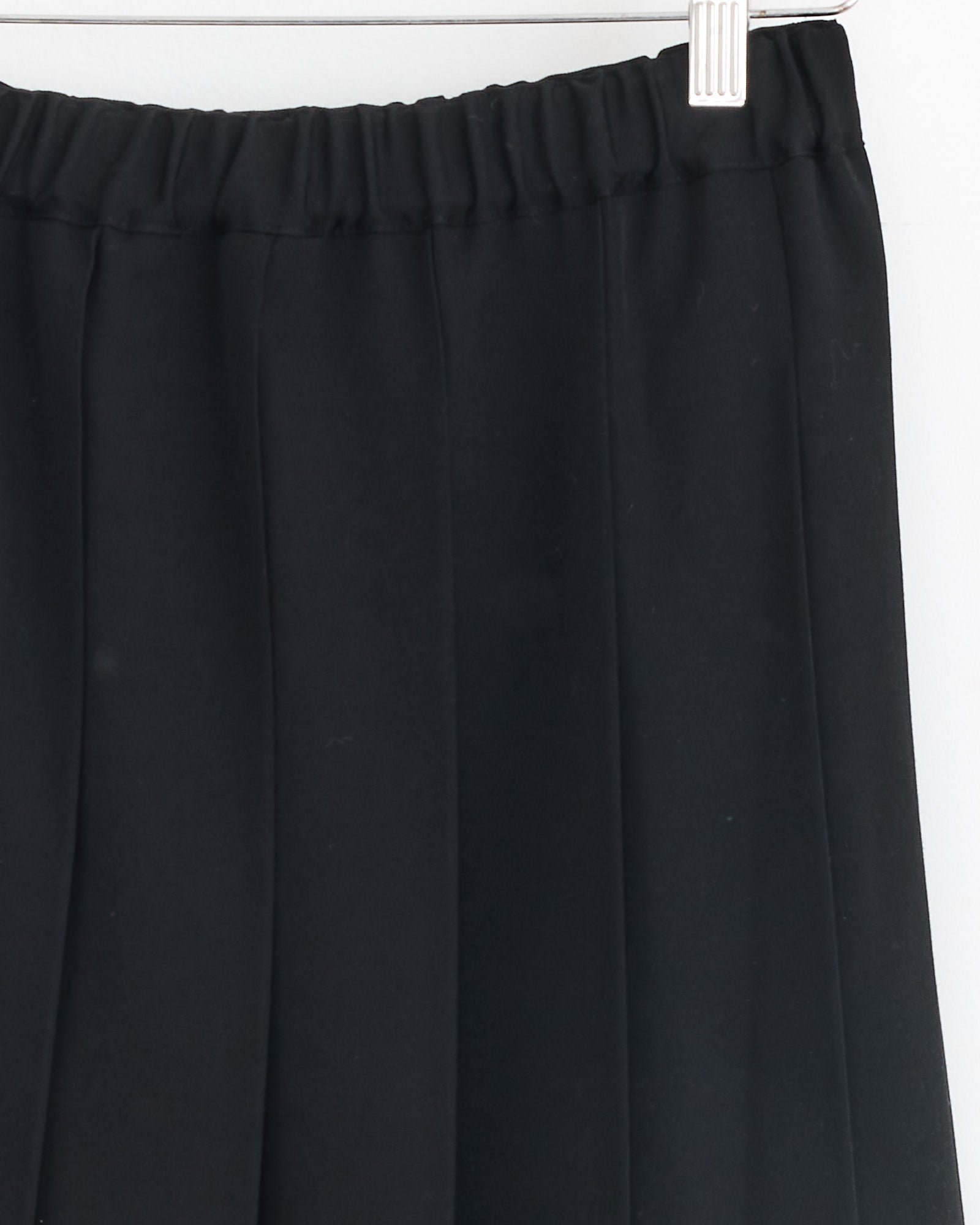 A close-up of the Comme Des Garçons Girl Pleated Skirt in Black, featuring tropical wool and an elastic waistband, made in Japan, shown on a white hanger against a white background.