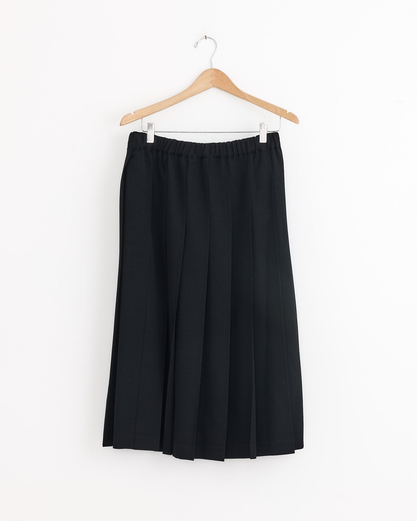The Pleated Skirt in Black by Comme Des Garçons Girl, made from tropical wool with an elastic waistband, hangs on a wooden hanger against a plain white wall.