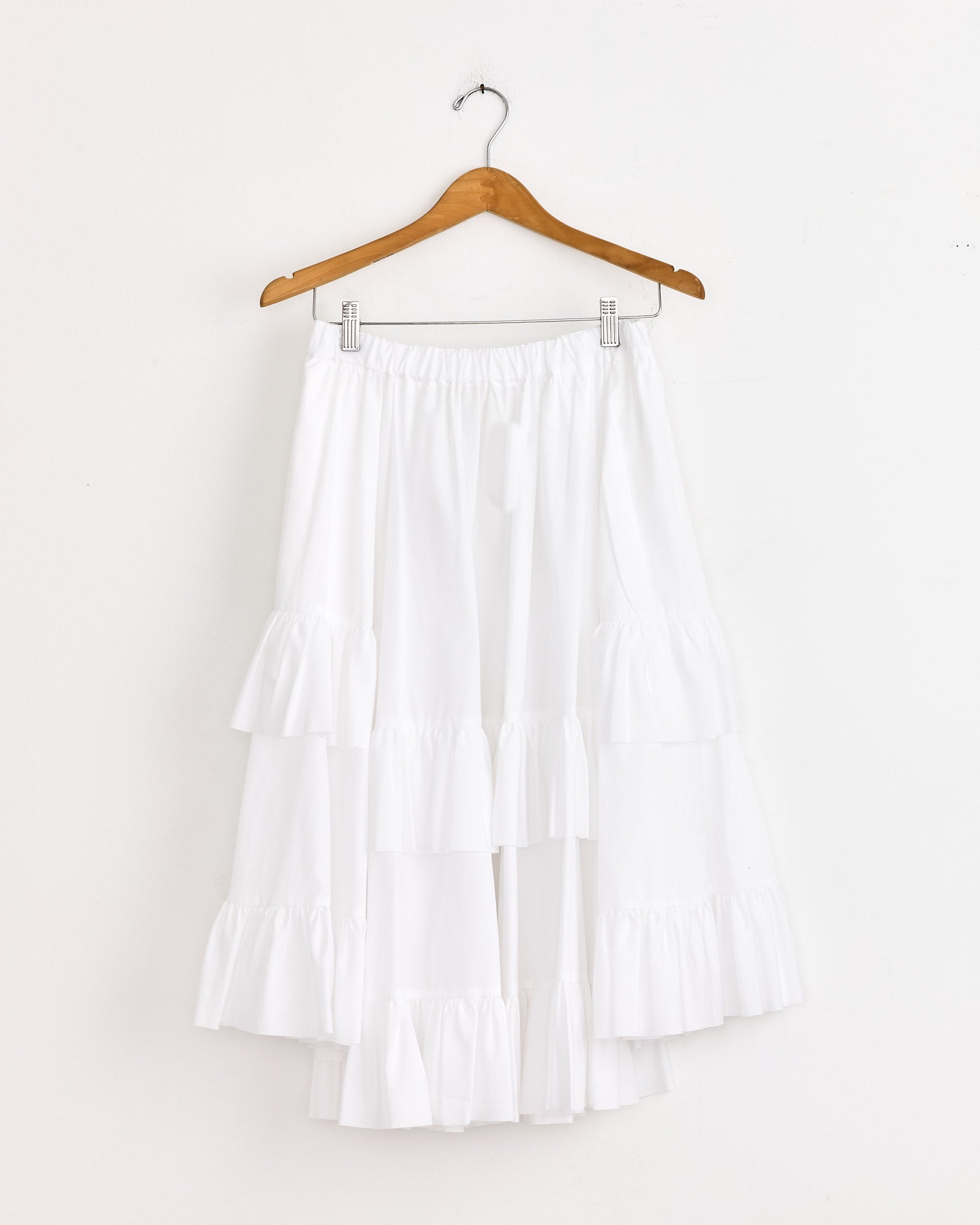 The Ruffled Skirt in White by Comme Des Garçons Girl, featuring layered ruffles, hangs elegantly on a wooden hanger against a plain white wall.