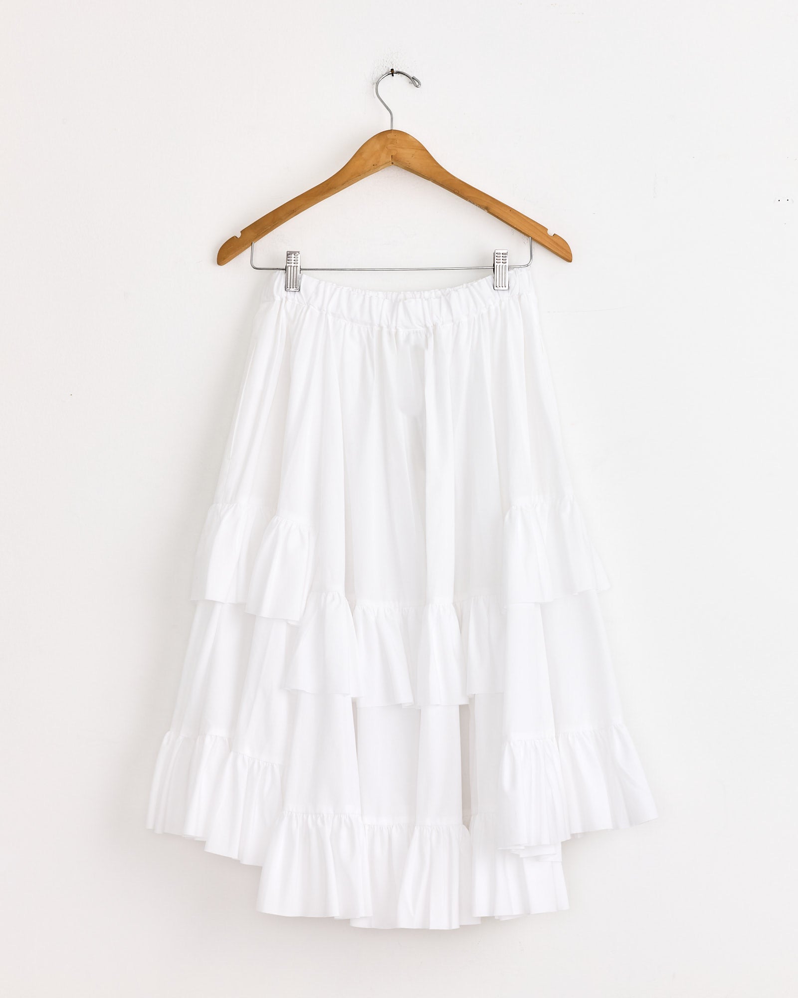 The Comme Des Garçons Girl Ruffled Skirt in White hangs on a wooden hanger against a plain wall, showcasing an elastic waistband and three tiered layers for a voluminous, flowy silhouette.