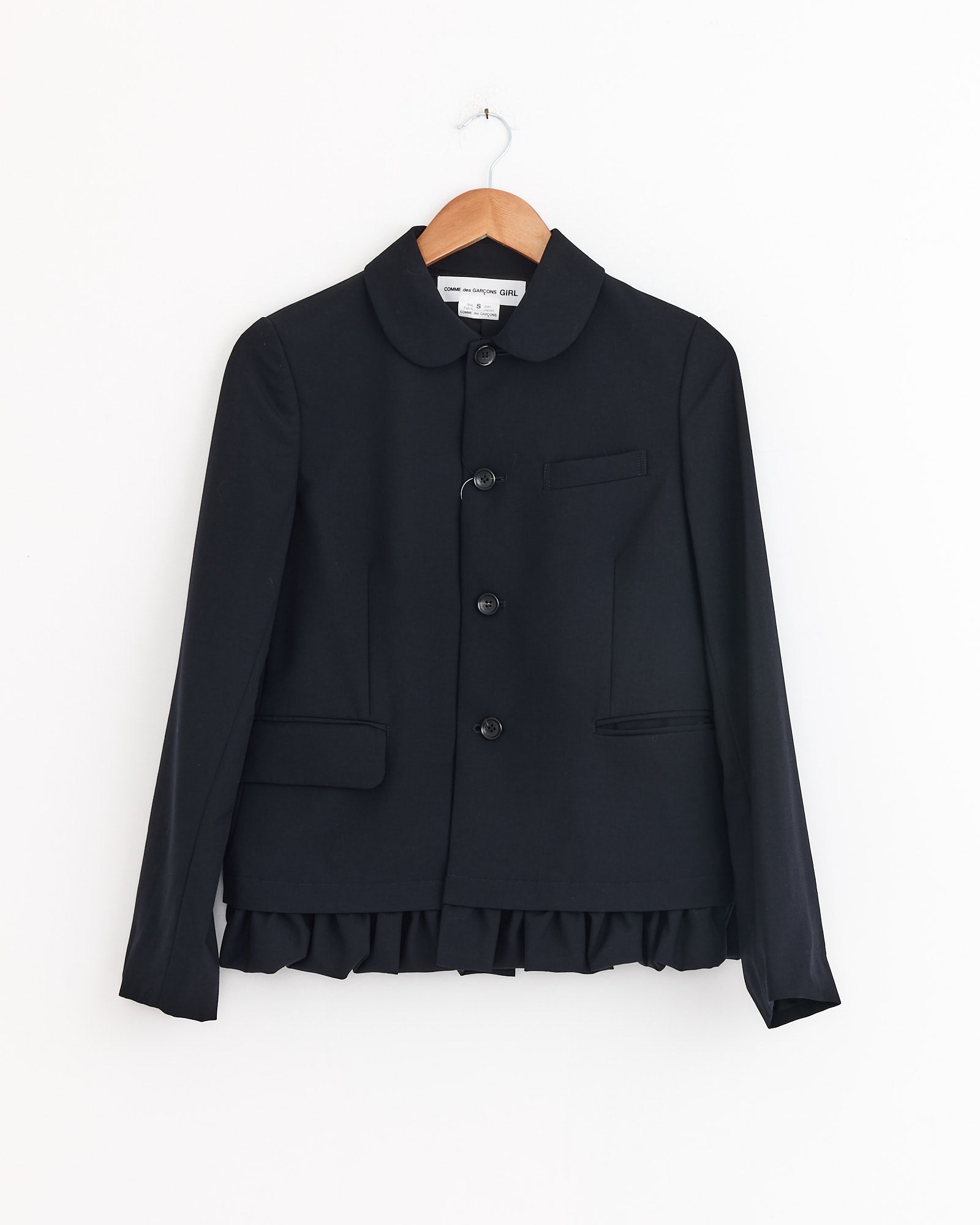The Comme Des Garçons Girl Peter Pan Jacket in Navy features long sleeves, a Peter Pan collar, three front buttons, two flap pockets, a chest pocket, and a ruffled hem. Displayed on a wooden hanger against a white background. Made in Japan.