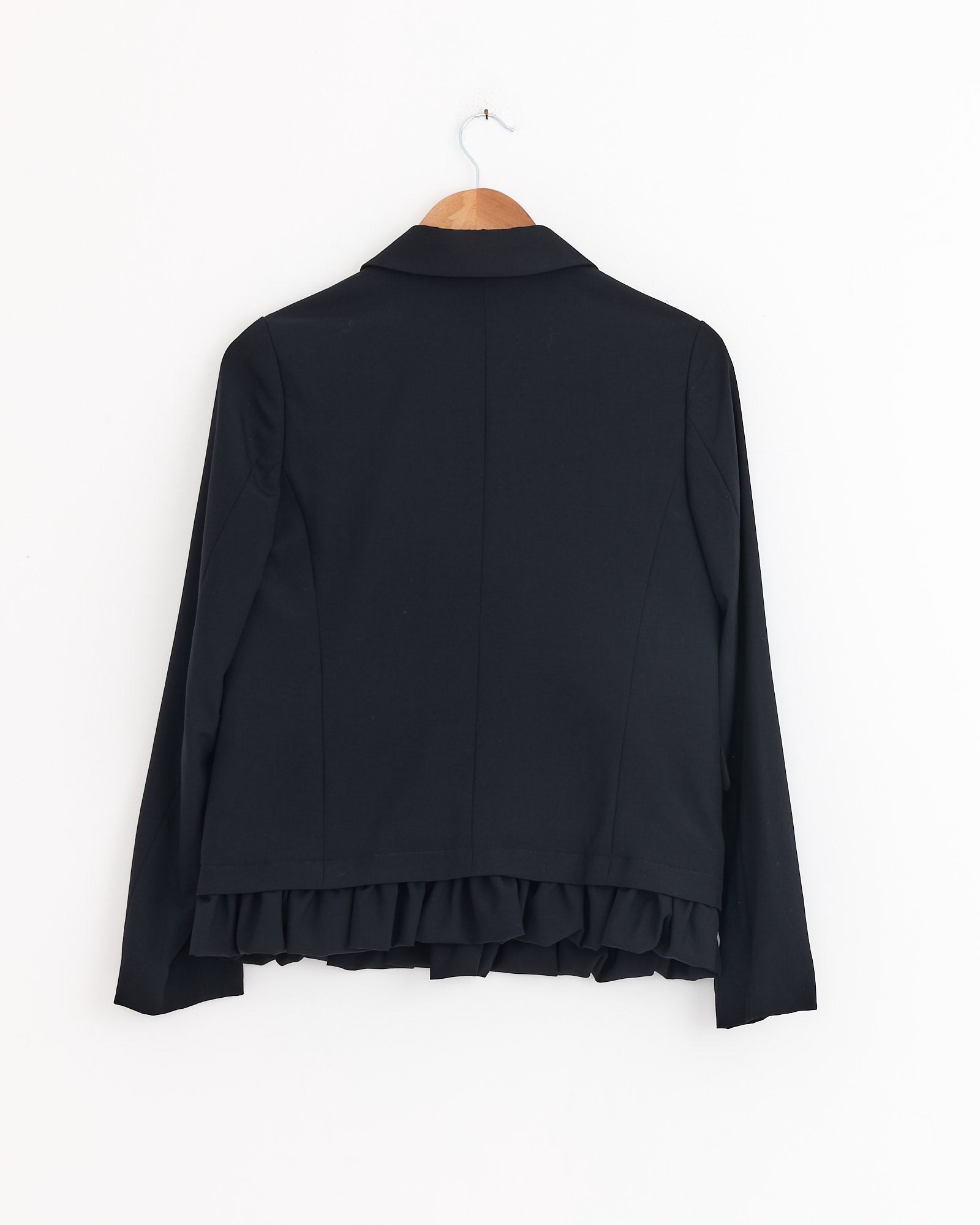 The Comme Des Garçons Girl Peter Pan Jacket in Navy, featuring a notched collar and ruffled hem, hangs on a wooden hanger against a white wall.