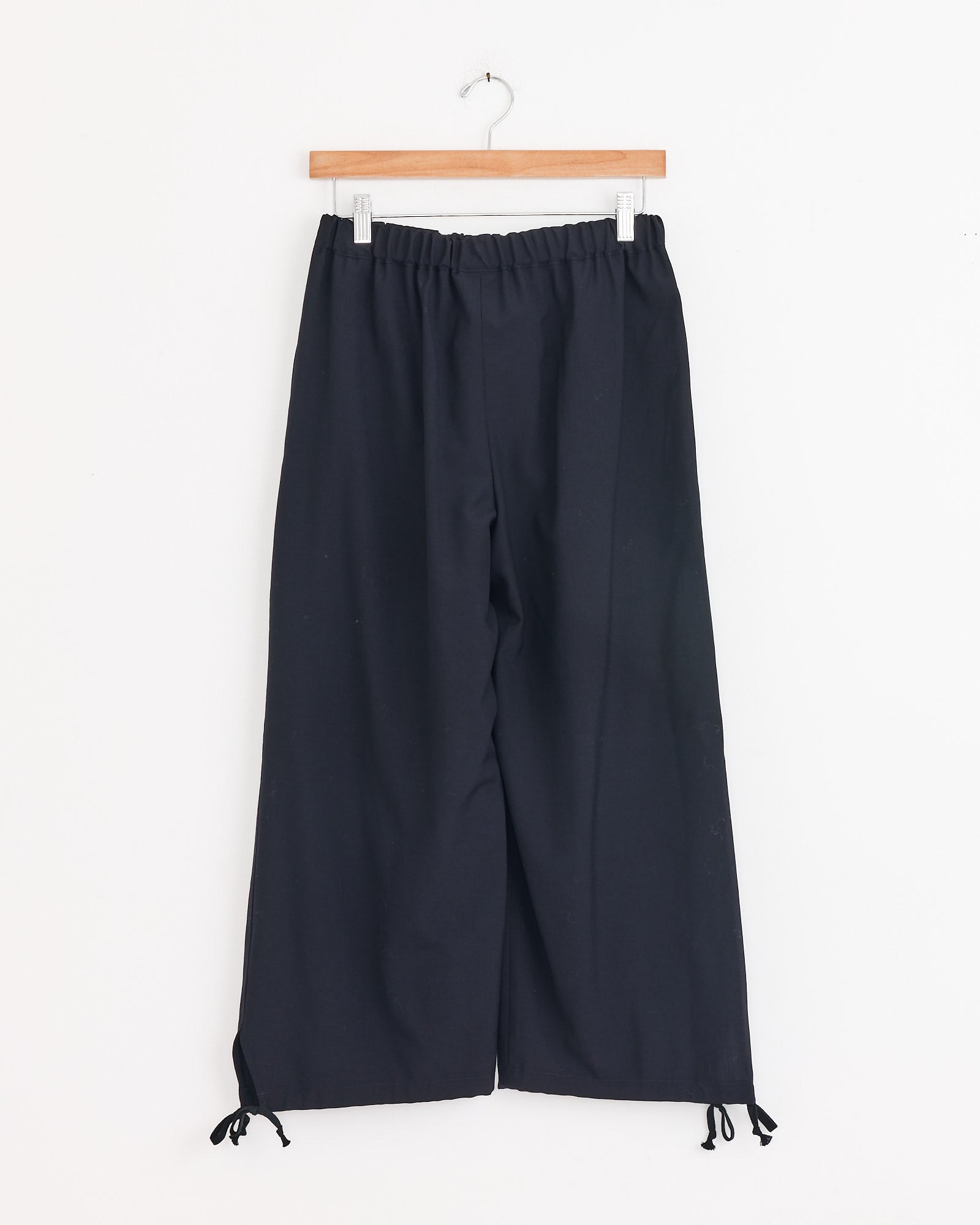 The Drawstring Cuff Pant in Navy by Comme Des Garçons Girl features wide legs, an elastic waistband, and adjustable ankle ties. Pictured on a wooden hanger against a plain white background, it showcases the brand's classic trouser silhouette.