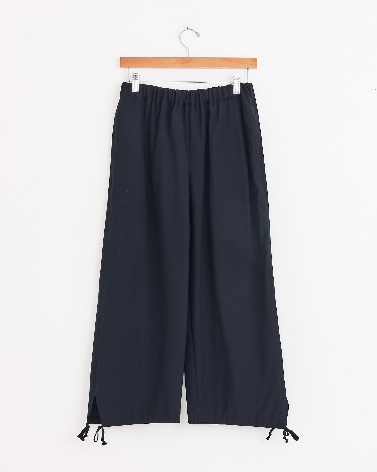 A pair of loose-fitting navy Drawstring Cuff Pants by Comme Des Garçons Girl hangs on a wooden hanger, featuring an elastic waistband and adjustable ties at the wide-legged hems, set against a plain white background.