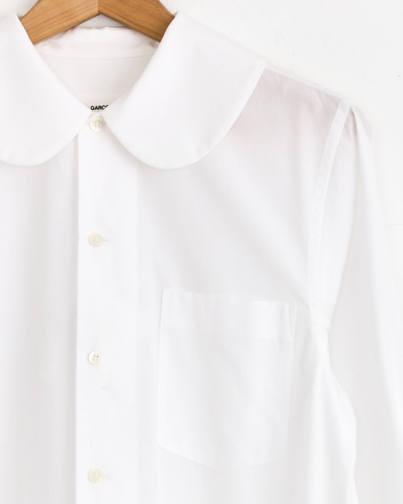 The Comme Des Garçons Girl Wide-Pleated Shirt Dress in white hangs on a wooden hanger against a plain white background.