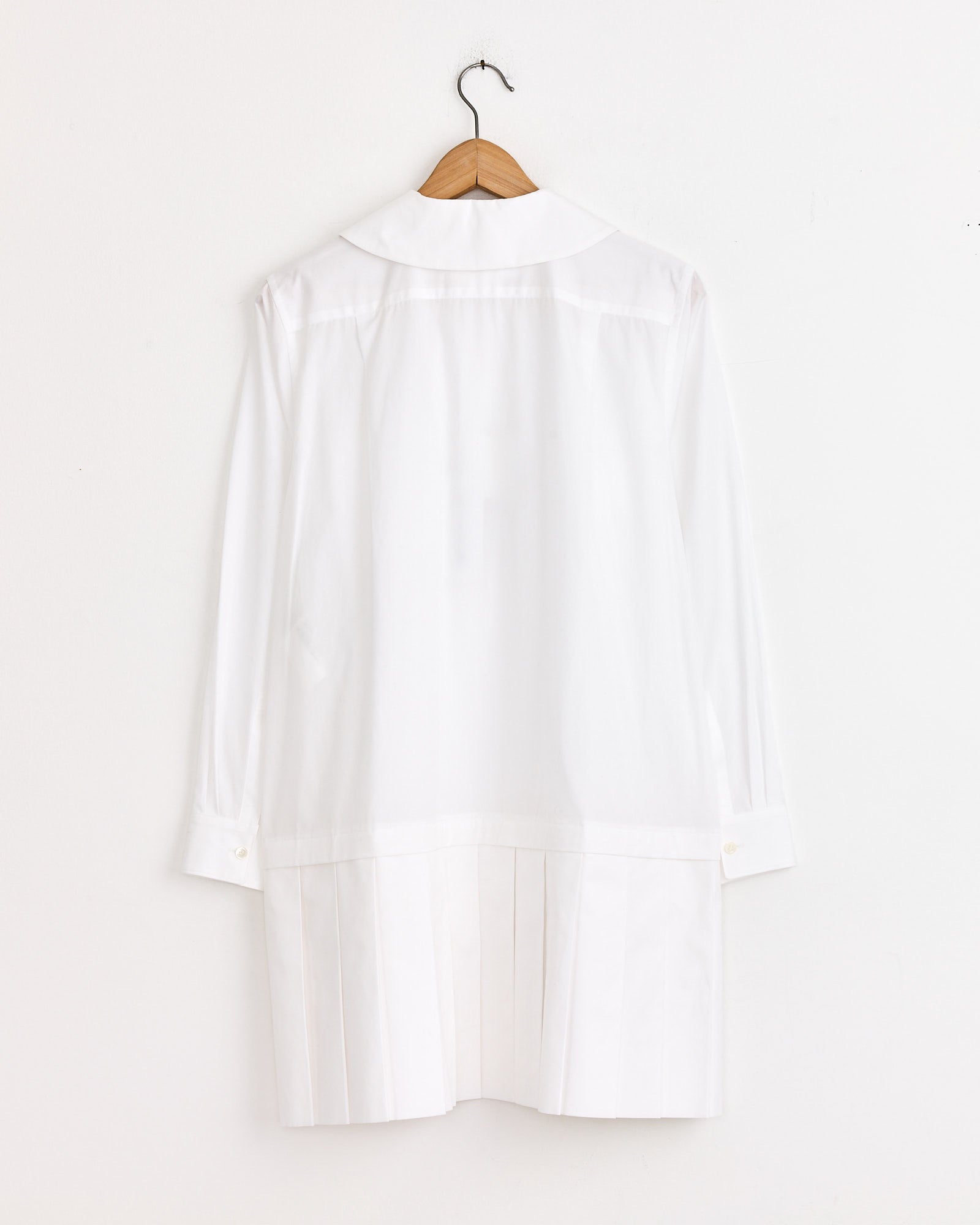 The Comme Des Garçons Girl Wide-Pleated Shirt Dress in White, featuring long sleeves and a round collar, hangs on a wooden hanger against a plain white wall.