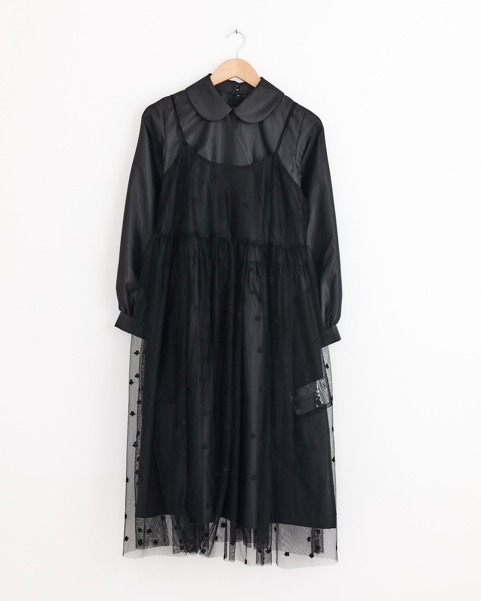 The Comme Des Garçons Girl Tulle Embroidered Overlay Dress in Black features long sheer sleeves, a Peter Pan collar, layered tulle skirt, and delicate flower embroidery, displayed on a wooden hanger against a white background.