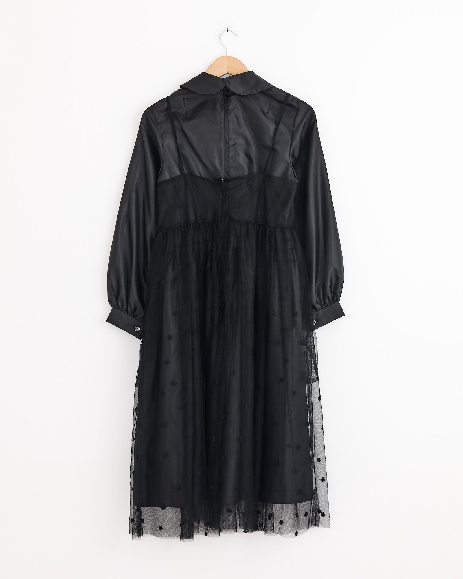 The Comme Des Garçons Girl Tulle Embroidered Overlay Dress in Black features long sleeves, a collared neckline, sheer tulle skirt overlay, and buttoned cuffs, displayed on a wooden hanger against a white background.